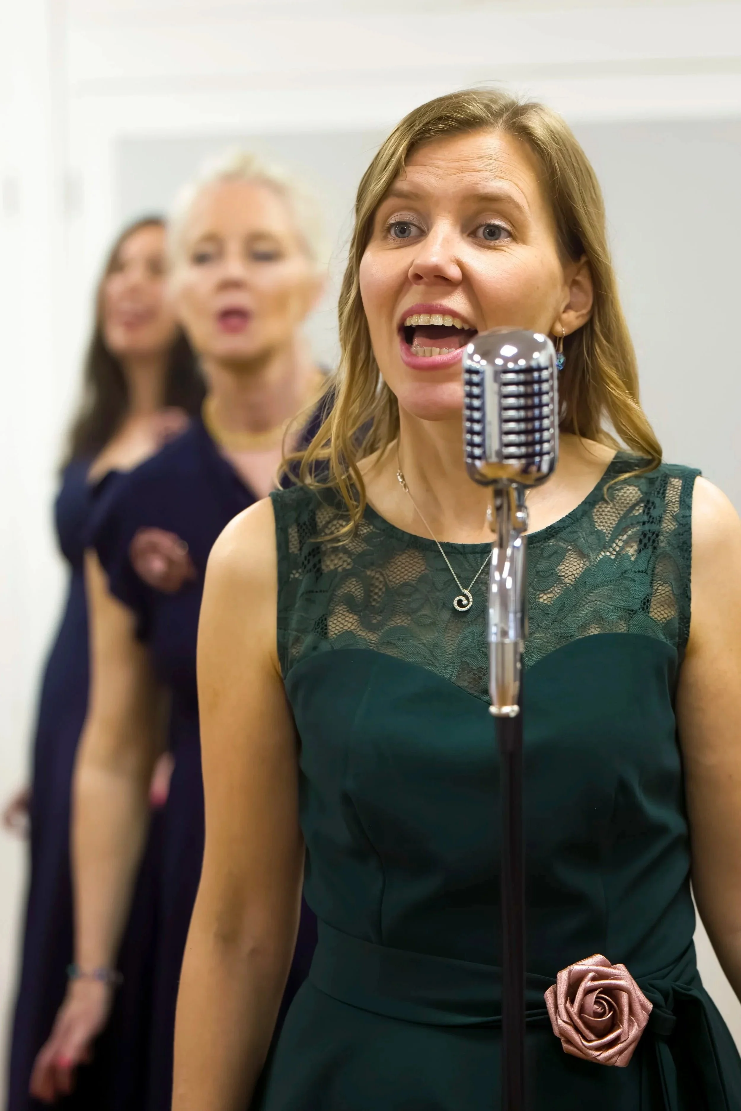 Woman from The Fifty choir singing into a vintage microphone, dressed in a dark green dress with a corsage, with two female choir members in the background, at an indoor performance in Southend-on-Sea.