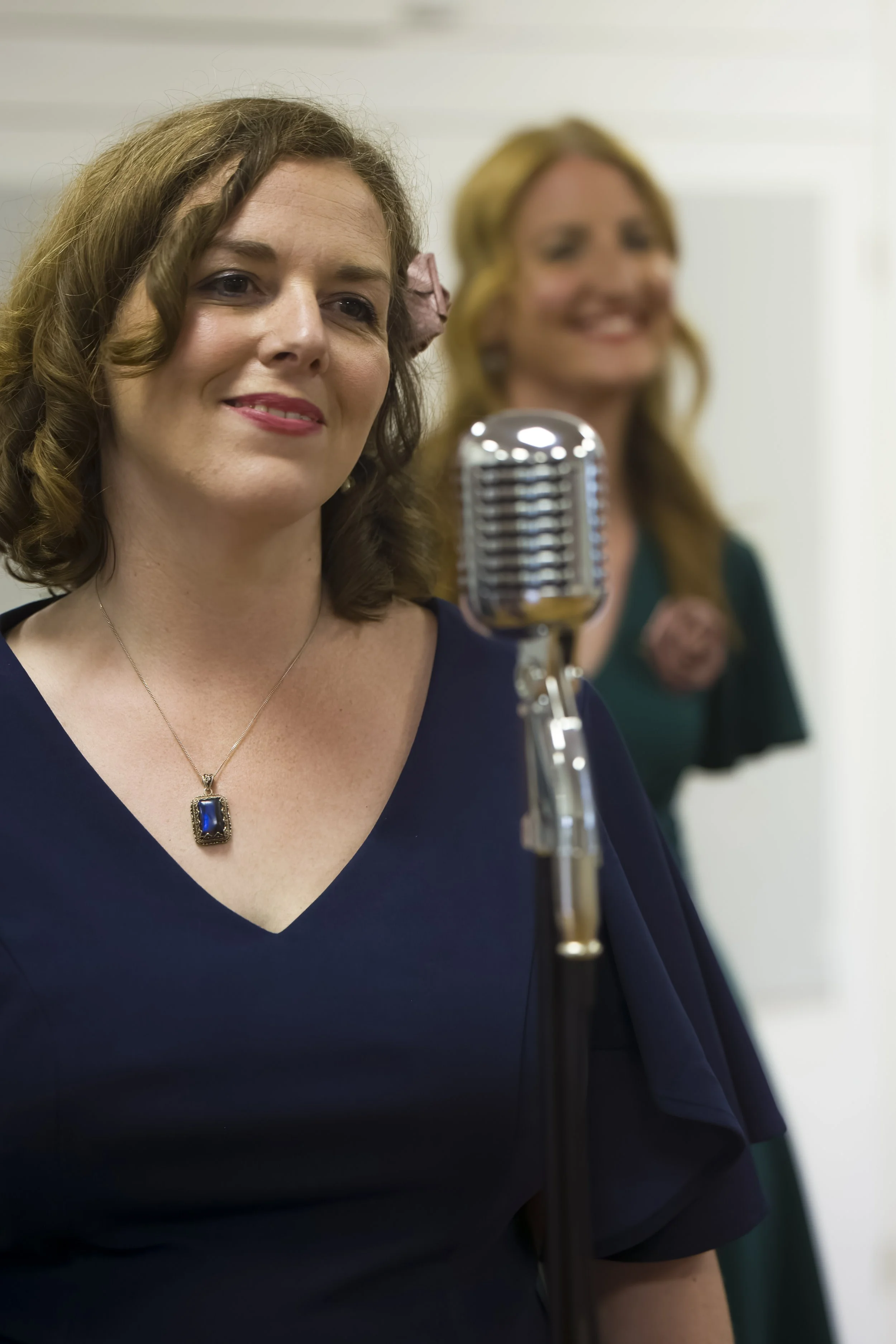 A woman with curly brown hair wearing a navy blue dress, a necklace with a large blue pendant, and a pink flower in her hair, smiling at a vintage microphone while another woman with wavy blonde hair and a green dress stands in the background.