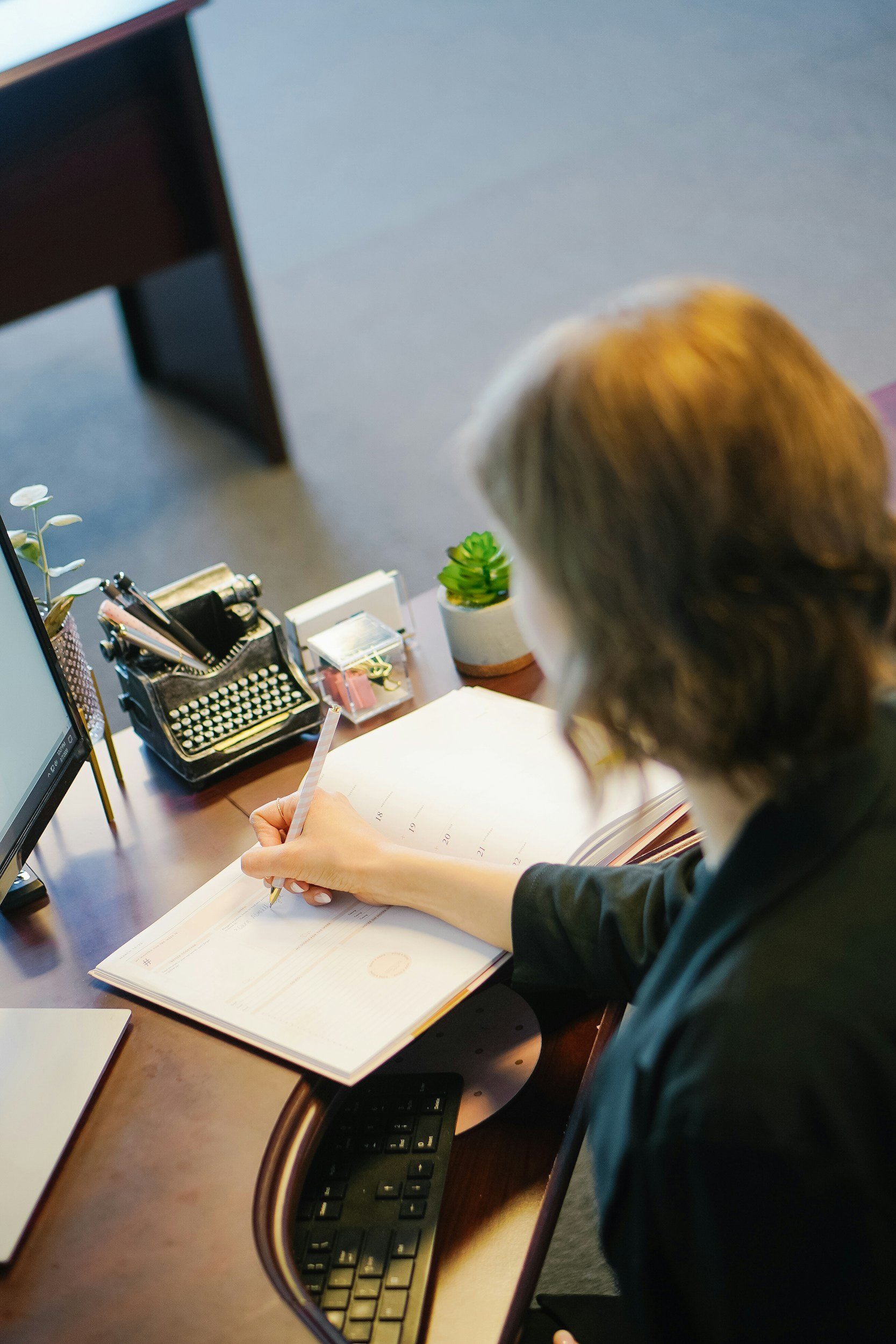 A woman with brown, shoulder-length hair, writing in a planner while sitting at a wooden desk. The desk has a calculator, a computer monitor, a black pen holder with pens, a small decorative plant, a white box, and a clear container with office supplies. There is also a small potted succulent plant. The background includes a gray wall and a small piece of dark furniture.