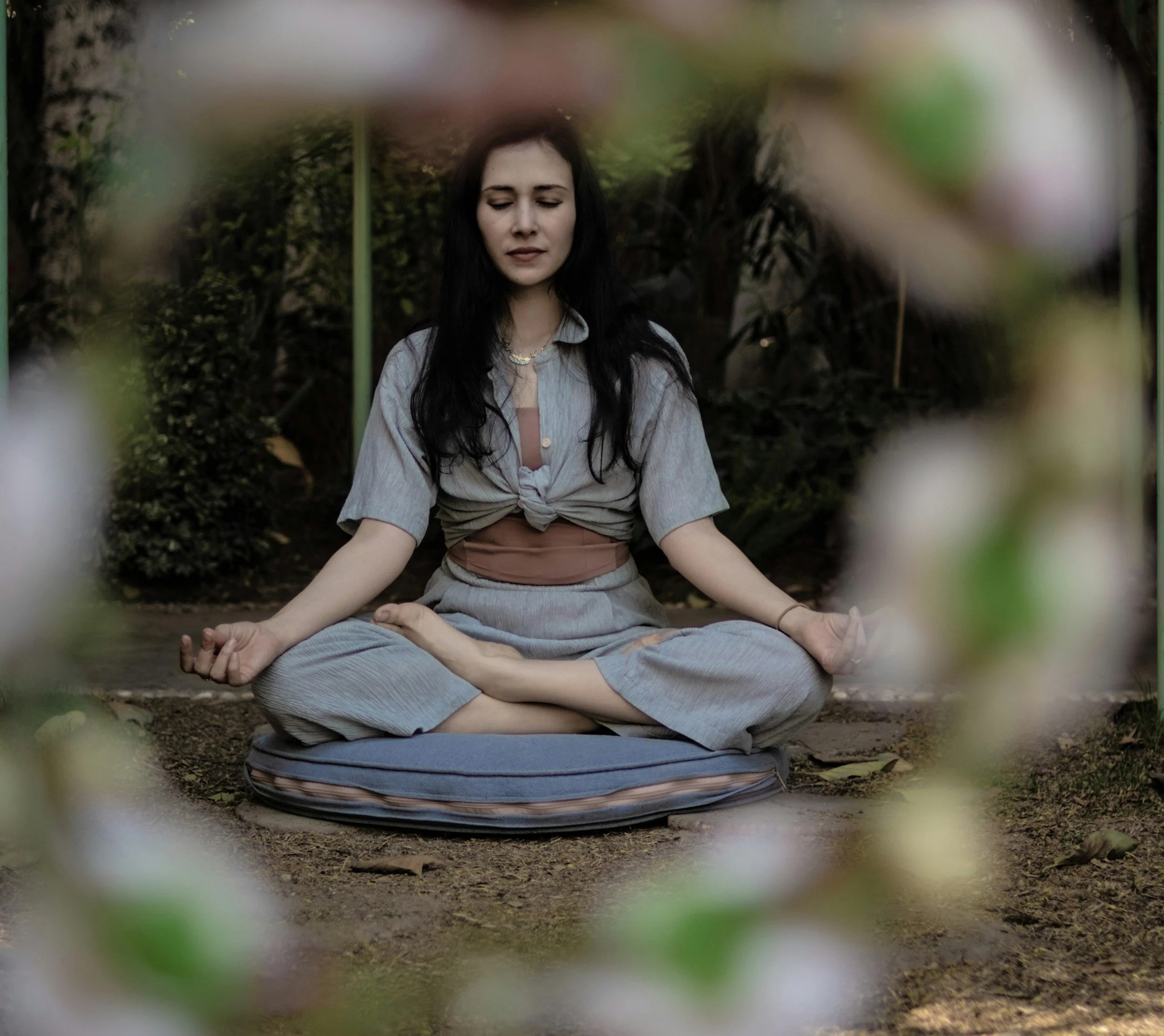 A woman practicing meditation outdoors, sitting cross-legged on a mat with eyes closed, surrounded by greenery and trees.