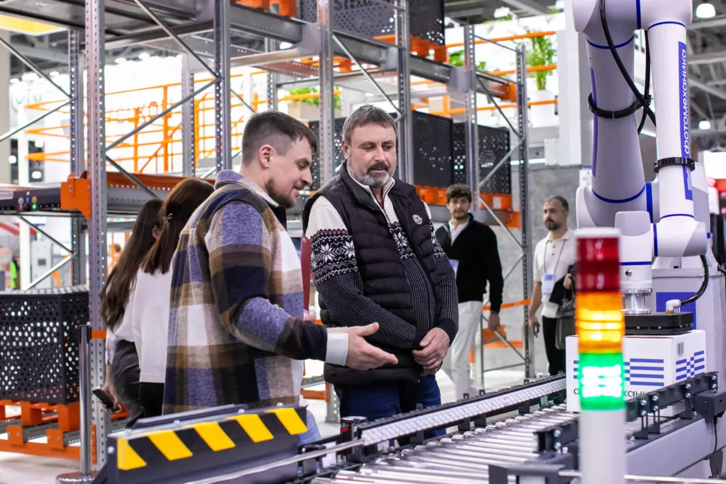 People observe a robotic arm and a colorful LED display in an industrial or research environment with metal shelves and electronic equipment in the background.