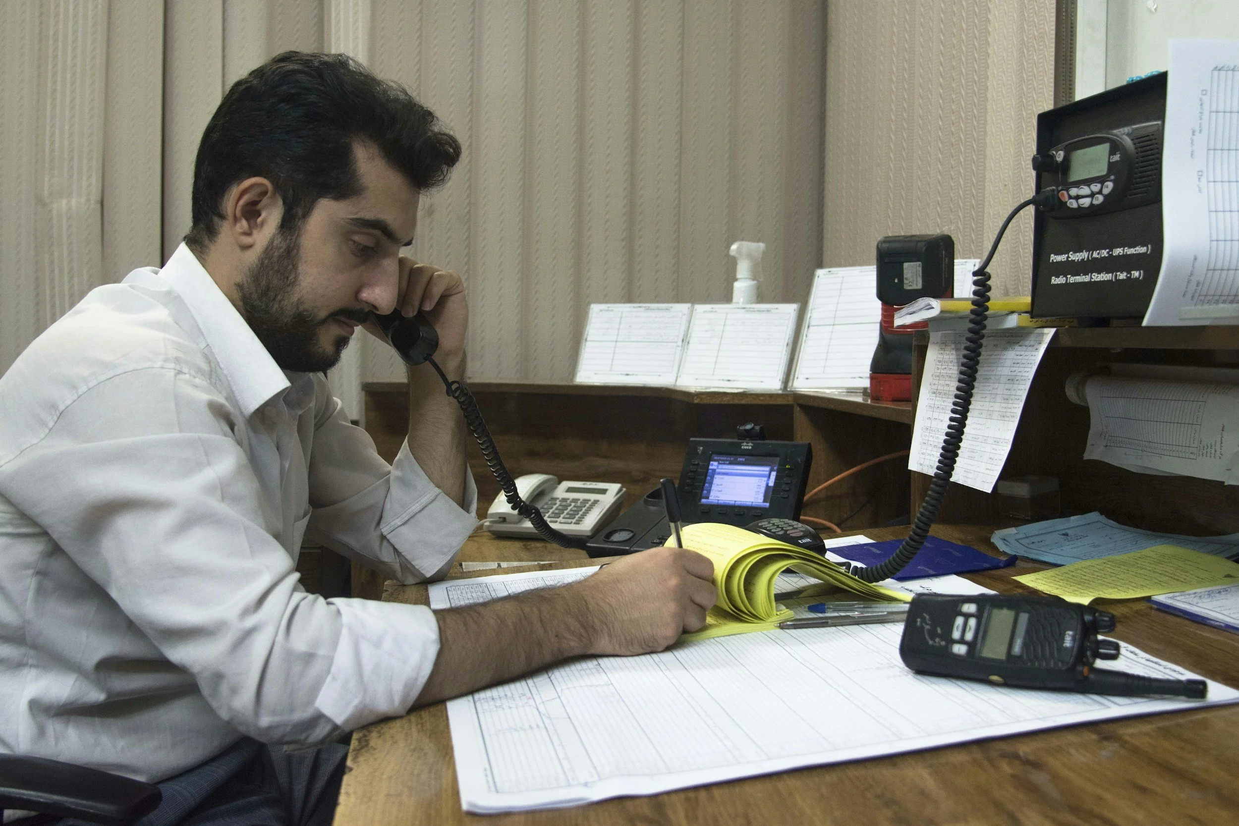 A man in a white shirt is sitting at a cluttered desk, talking on a corded telephone, writing on a yellow notepad, with various papers, a computer monitor, and office supplies around him.