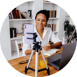 A woman sitting at a desk with a laptop, appearing to record or stream using a camera on a tripod, in a room with bookshelves in the background.