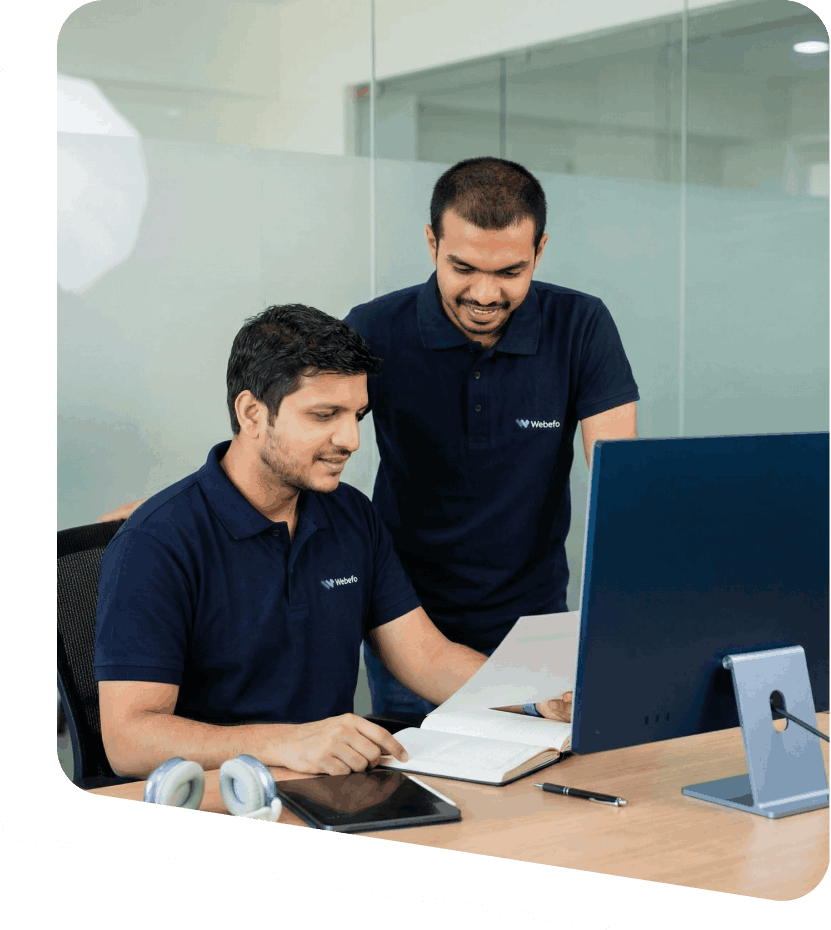 Two men in dark blue polo shirts working at a desk with a computer, notebook, tablet, pen, and headphones, in a modern office setting.
