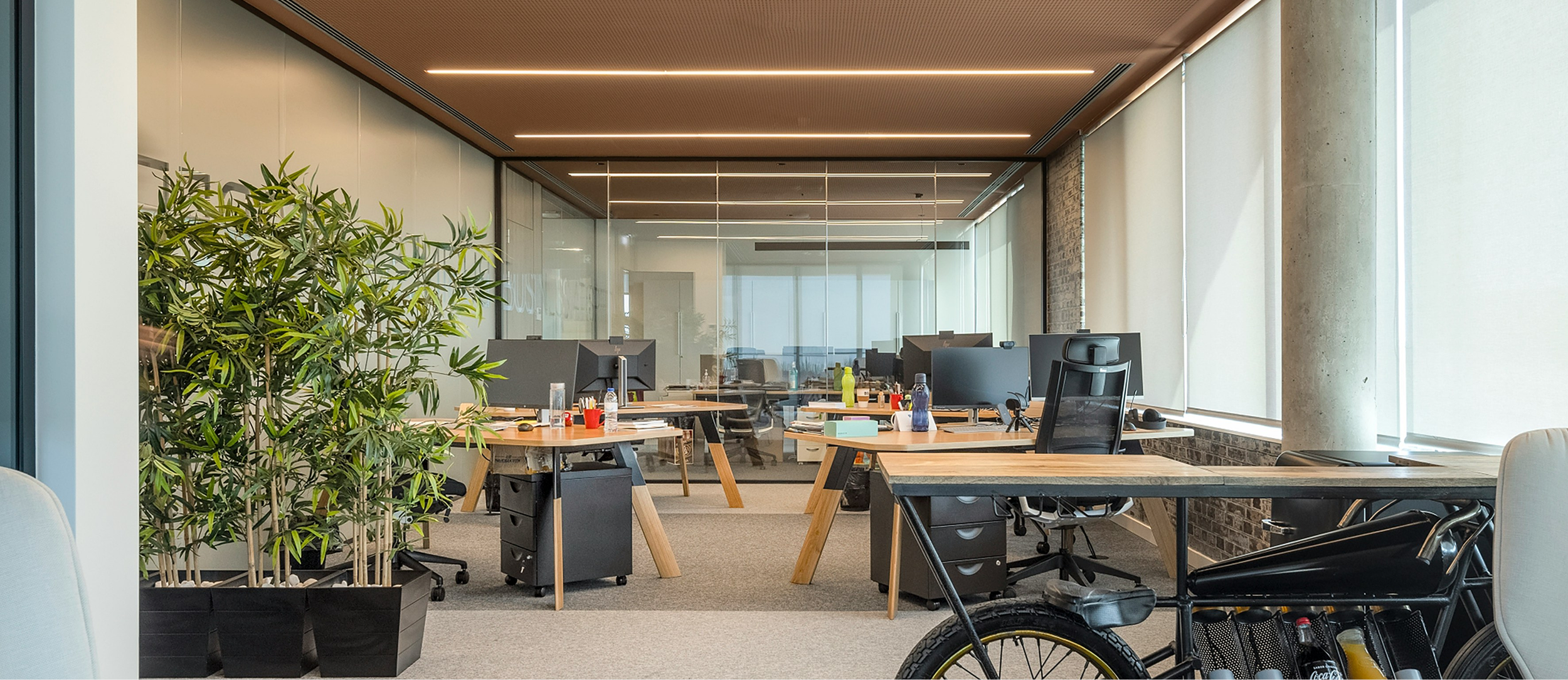 Modern open-plan office with desks, computers, chairs, and a bicycle, separated by glass walls, with large windows allowing natural light.
