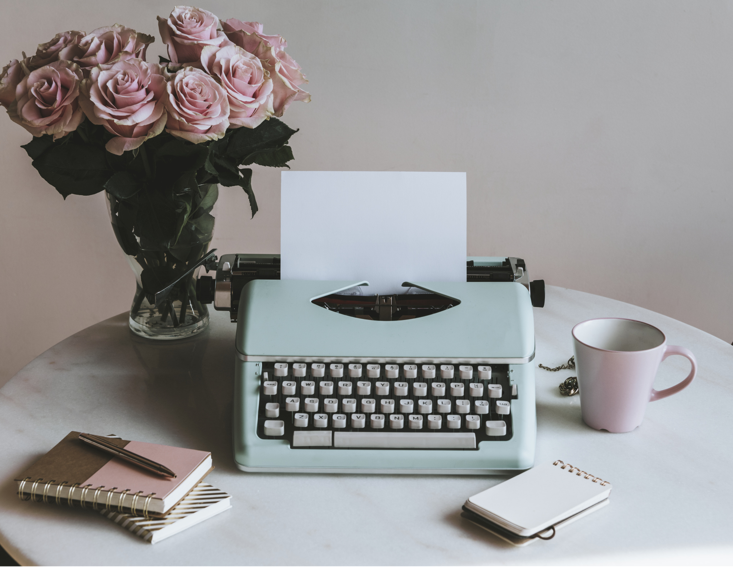 Image of 'old fashioned' typewriter in duck egg blue with paper inserted and typing that says "Dear Annie,". Theres a vase on the table with pink roses and a pink coffee cup and a couple of note books and pens