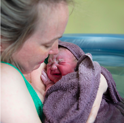 She did it! New mama in the birthing pool cuddles her newborn to her chest. Wrapped in a towel, the baby is pink and adapting to life 'outside'. This amazing Mama looks down adoringly at her little one.