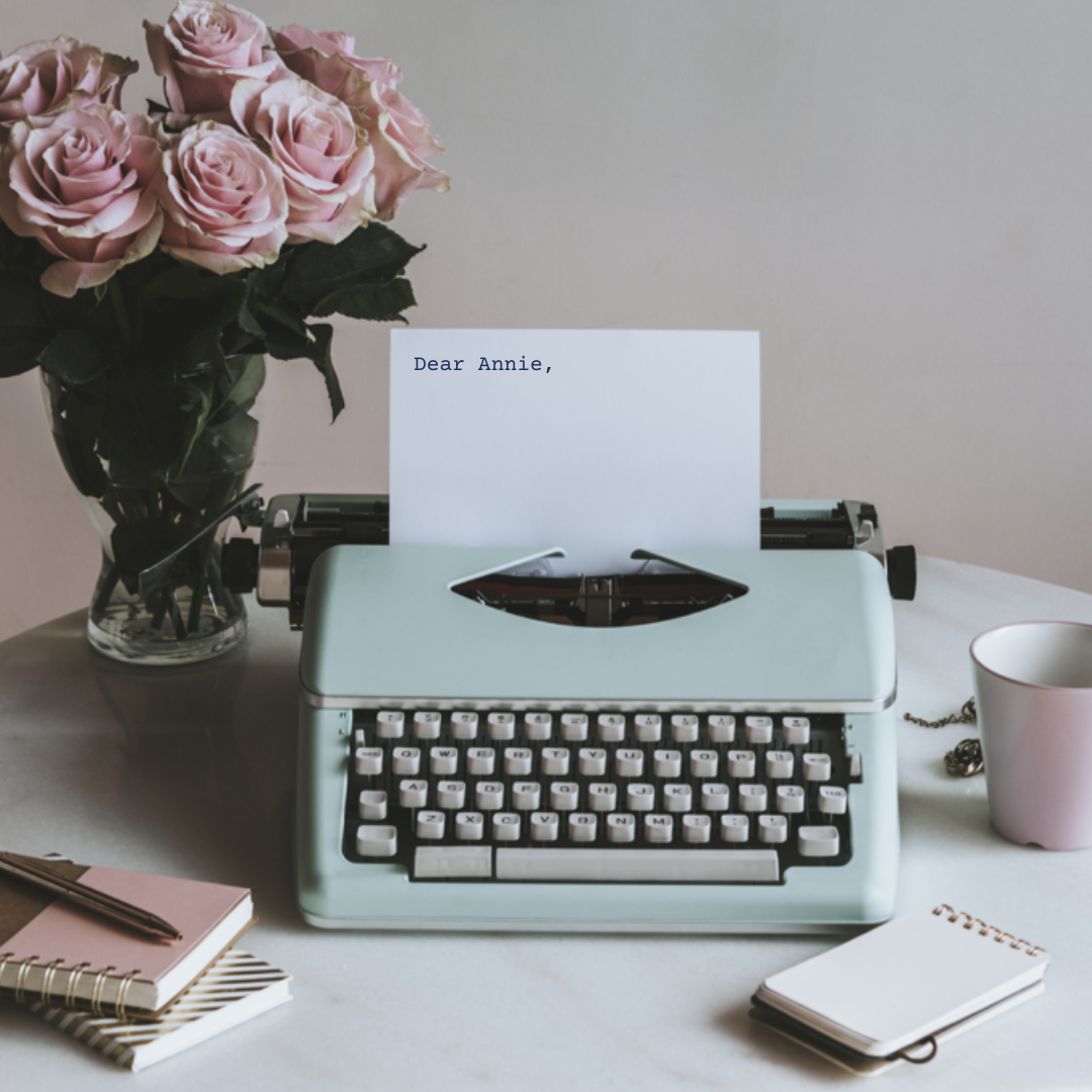 Old fashioned typewriter on desk with flowers, notebooks and a cup of coffee scattered on the desk. The paper fed into the typewriter reads "Dear Annie"