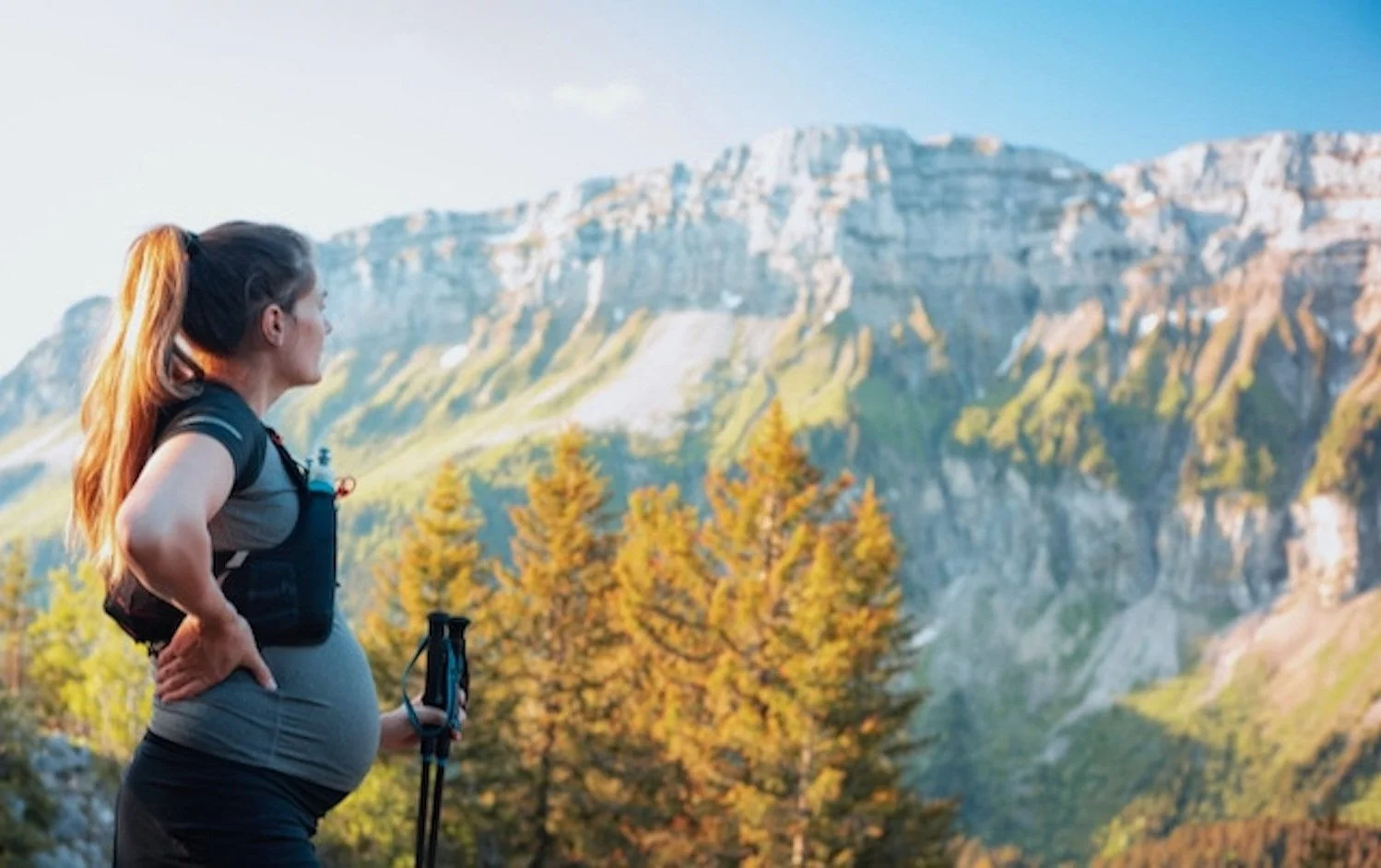 Pregnant woman stands in countryside looking at the mountains across the valley. She holds walking poles as though having climbed to this viewpoint. She appears relaxed and confident in her surroundings.