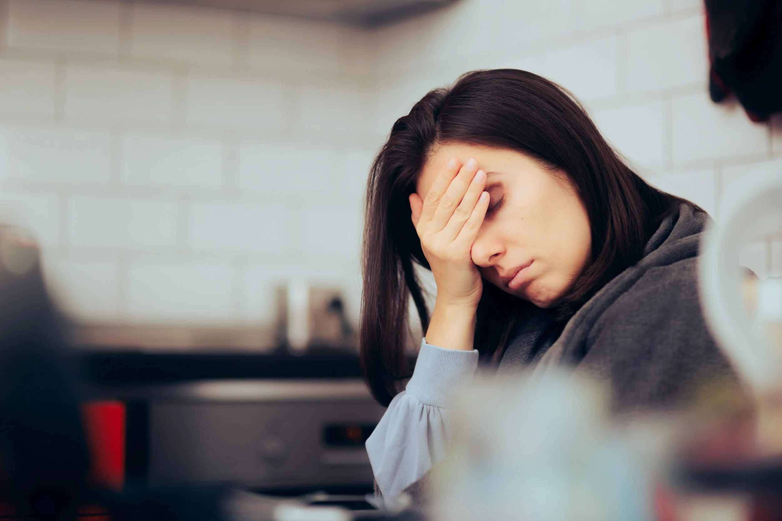 Dark haired young woman sits in her kitchen with her head in her right hand. Her eyes are closed and her face looks sad as though deep in thought and anxiety