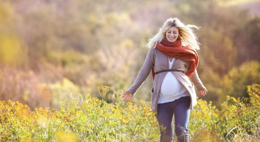 Blond haired pregnant woman walks towards camera within nature. Her hand brushes through the top of some yellow flowers. SHe smiles and appears confident and happy. 