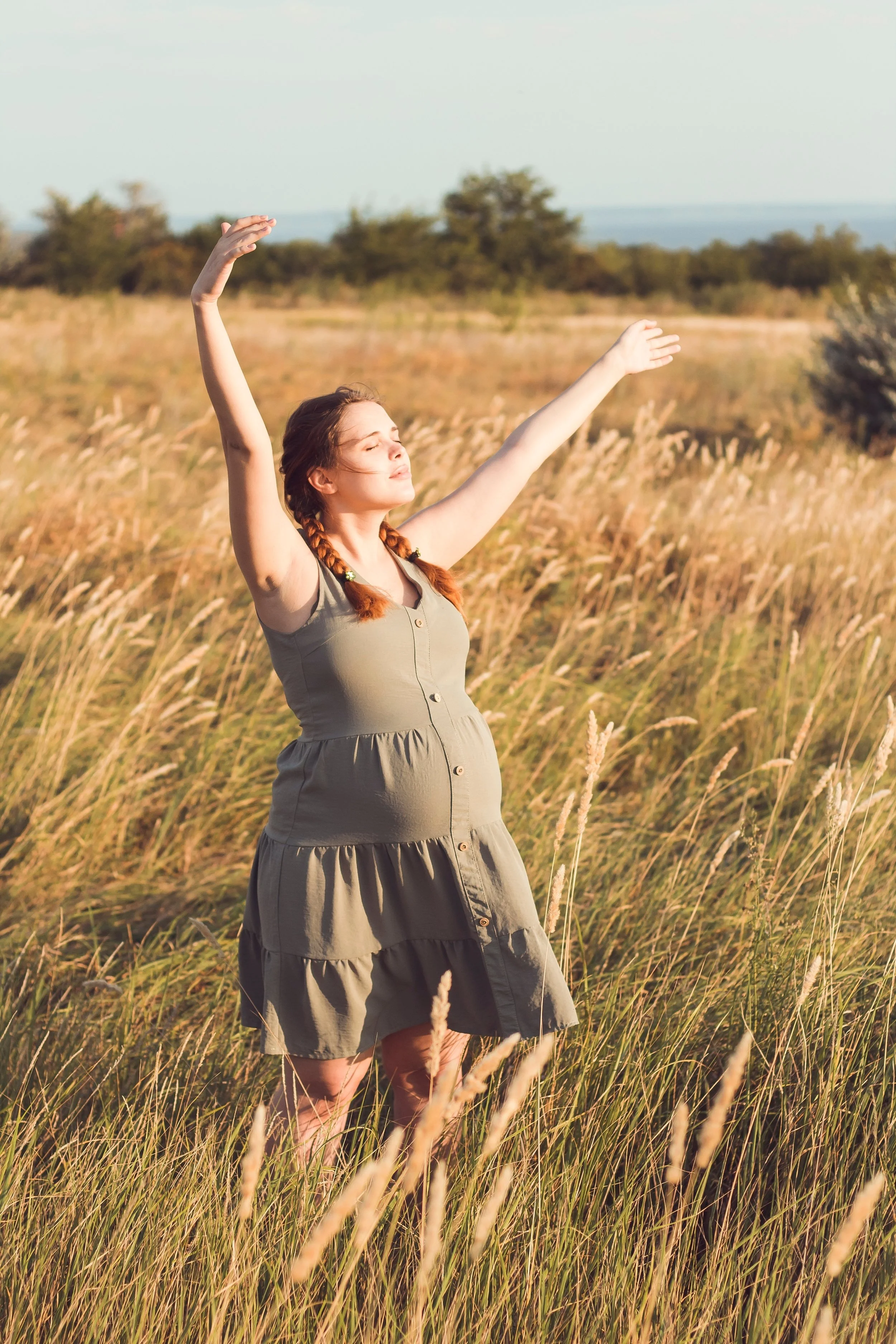 A pregnant woman dressed in a khaki dress stands in a field with long grasses. She faces towards the sun and her eyes are closed and her arms are outstretched as if welcoming the rays of light or maybe performing some dance moves.