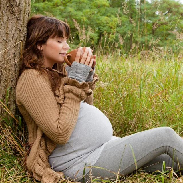 Dark haired pregnant woman rests against a tree with a mug of tea in her hands. She appears relaxed and ready for what comes next