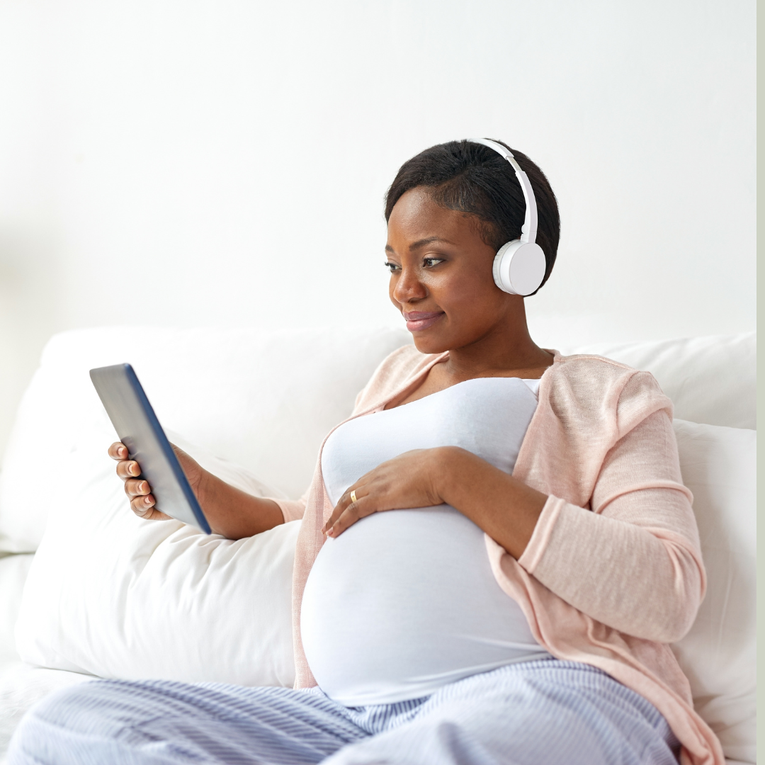 Coloured pregnant woman reclines with head phones on, an ipad in her hand. She is planning her birth journey and appears confident and totally at ease with whats happening. 
