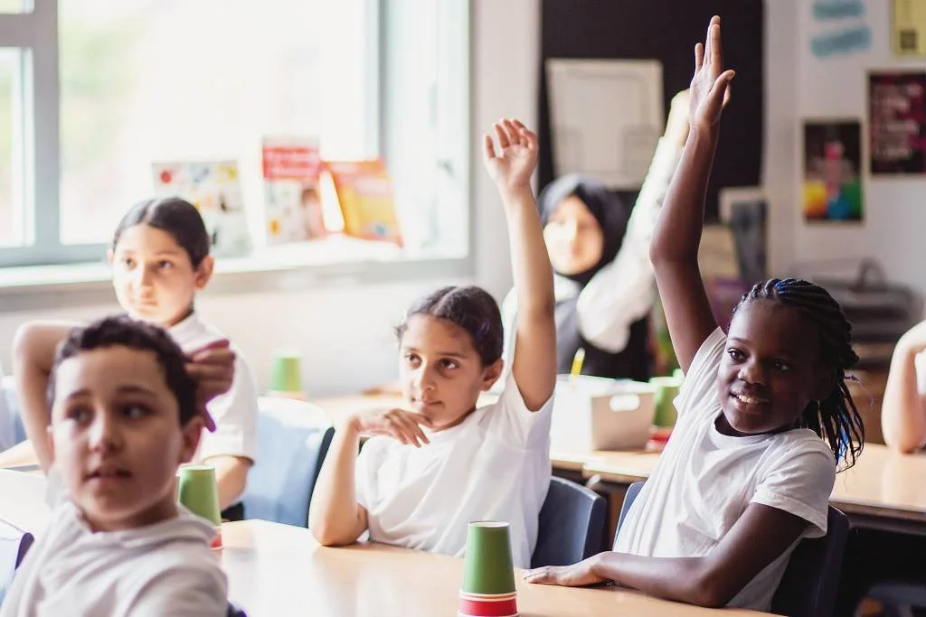 Classroom with students, some raising hands, and desks with cups, visible window, and educational posters.
