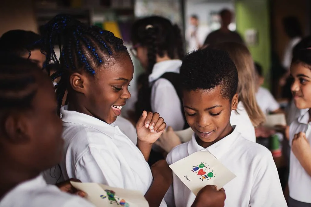 Children in white shirts smiling and looking at colorful holiday cards in a classroom