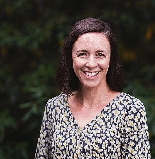 A woman with shoulder-length brown hair smiling outdoors with green foliage in the background, wearing a patterned blouse.