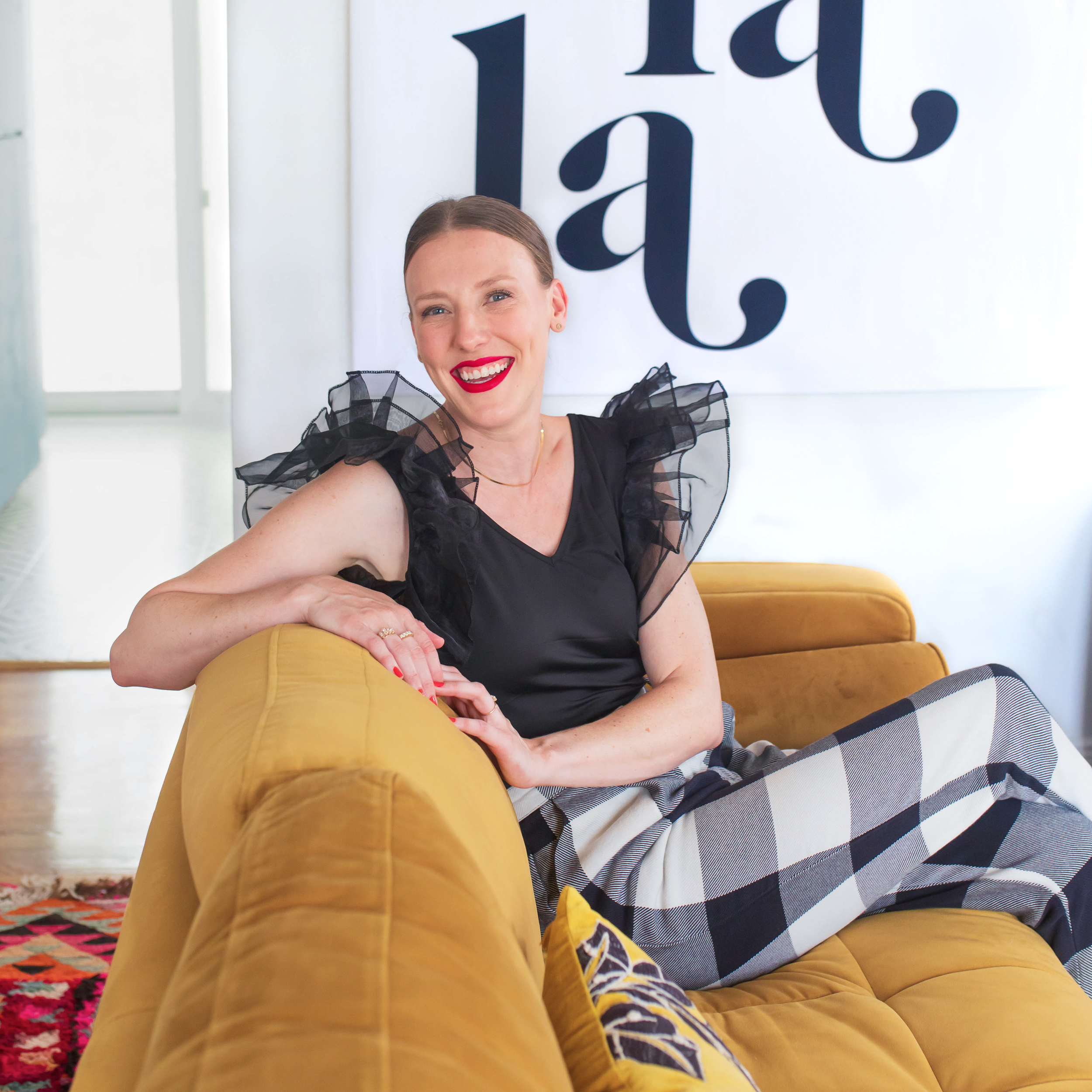 A woman with red lipstick and a black top with ruffled sleeves, sitting on a mustard-colored sofa, smiling at the camera in a bright room with a white background and large black typography.