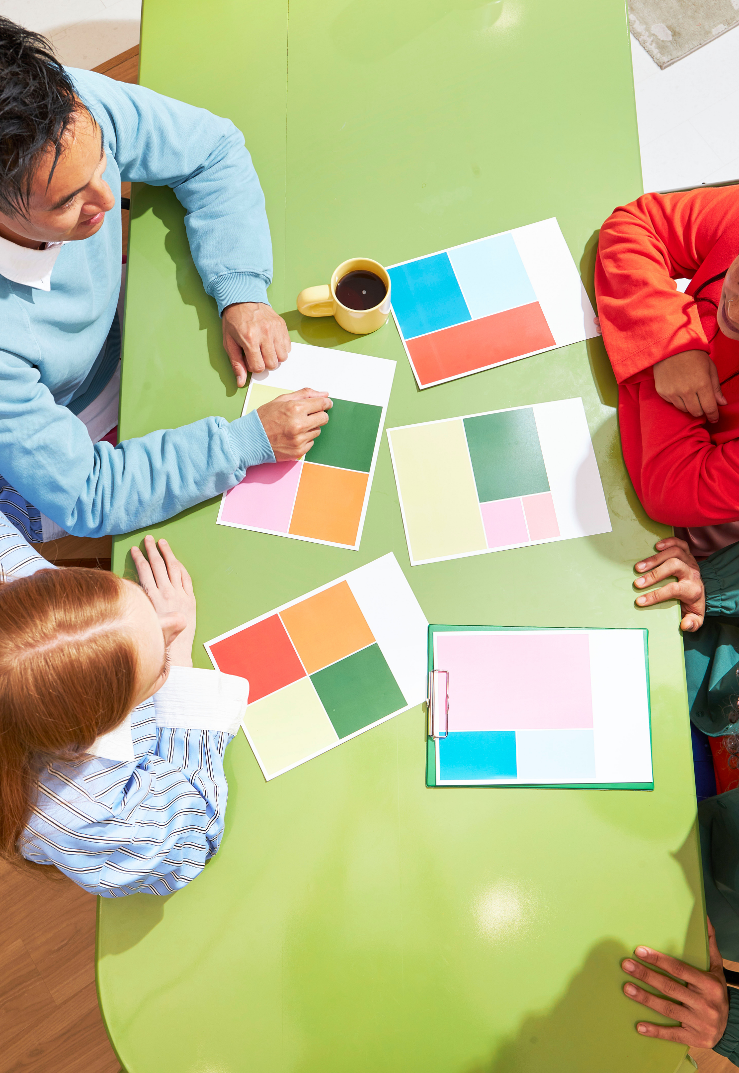 Three people sitting at a lime green table with color swatch cards, a yellow mug of coffee, and a clipboard, arranging color samples.