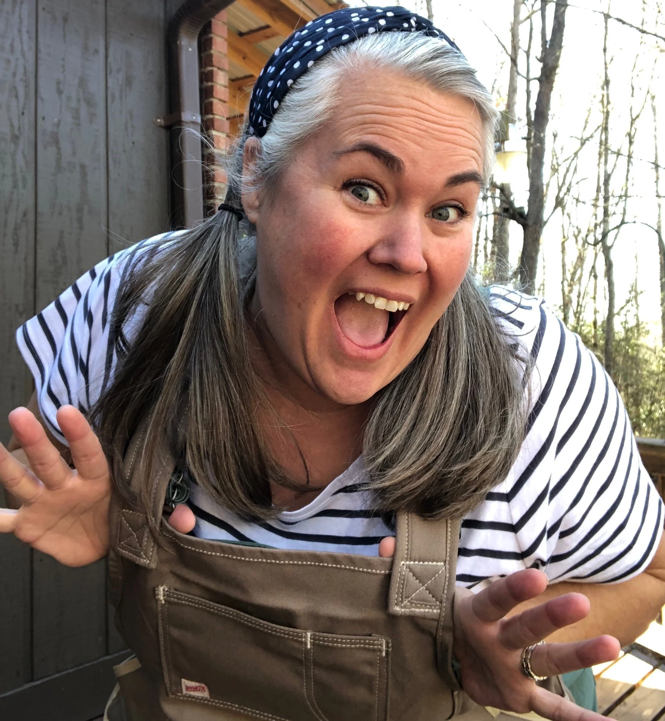 Joyful woman with gray hair, wearing a striped shirt, beige apron, and polka-dot headband, posing playfully outdoors.