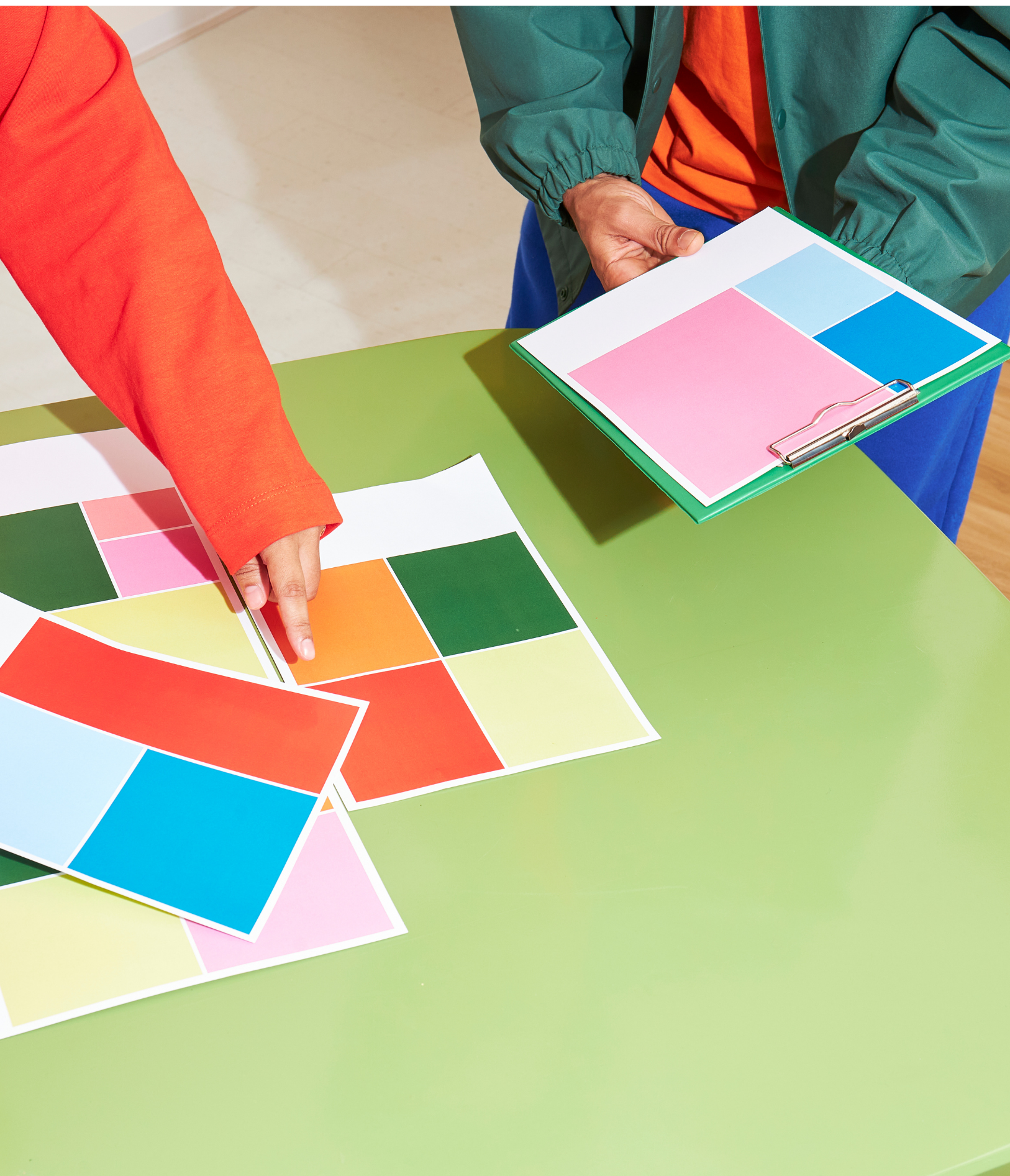 Two people are collaborating on color swatches and color palette samples laid out on a green table, with a clipboard holding additional color samples visible.