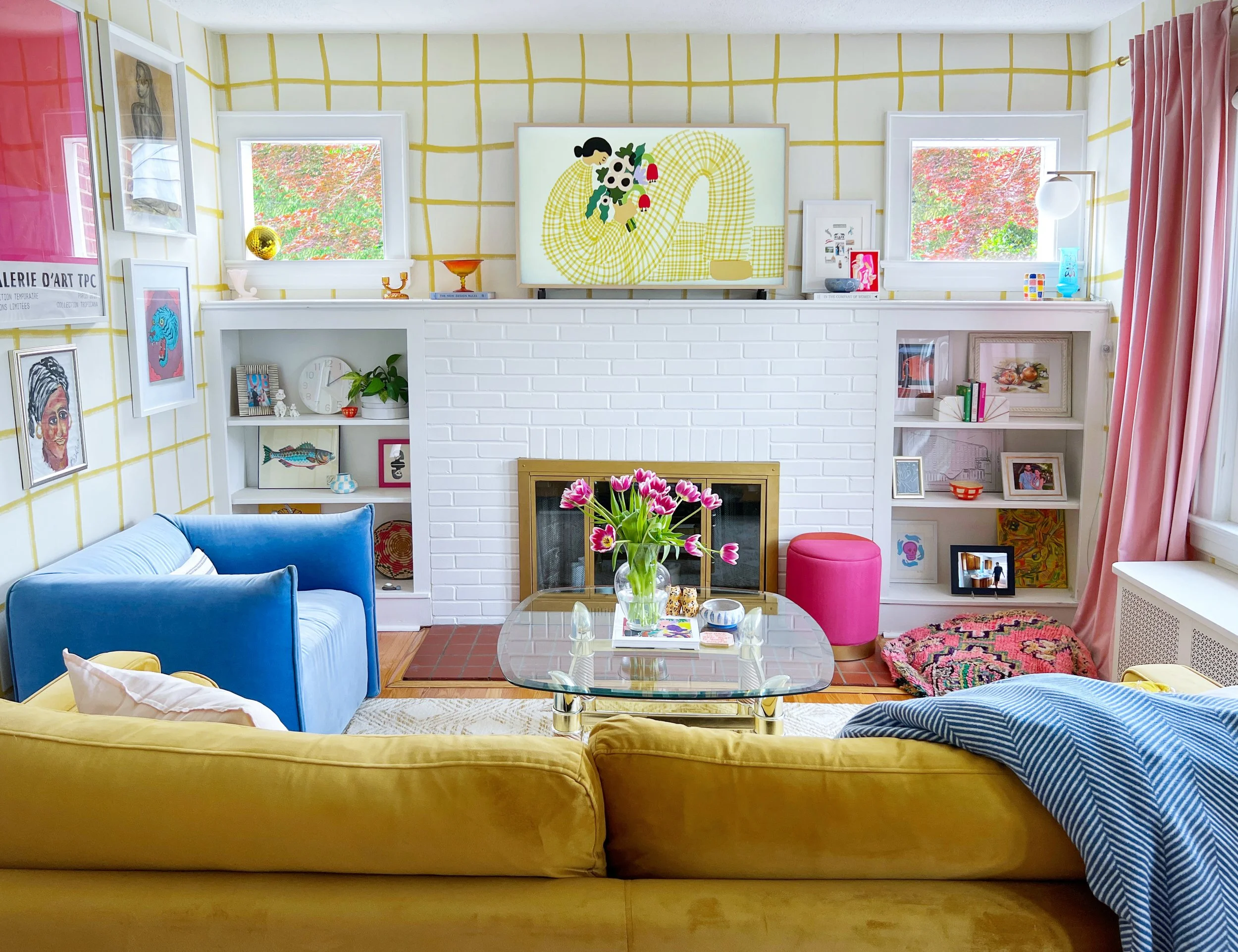 Colorful living room with yellow, pink, and blue furniture, white brick fireplace, glass coffee table with flowers, bookshelves with art and decor, and art on the walls.
