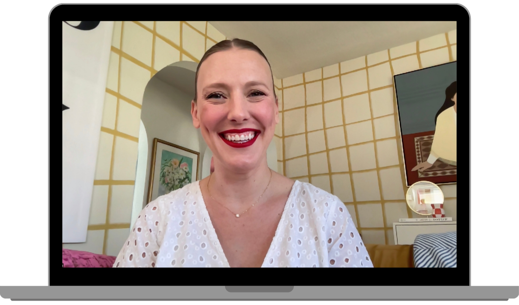 A woman with short hair wearing a white eyelet blouse and red lipstick, smiling at the camera, in a room with yellow checkered wallpaper, a framed picture of flowers on the wall, and a painting of a woman in a yellow dress on the right.