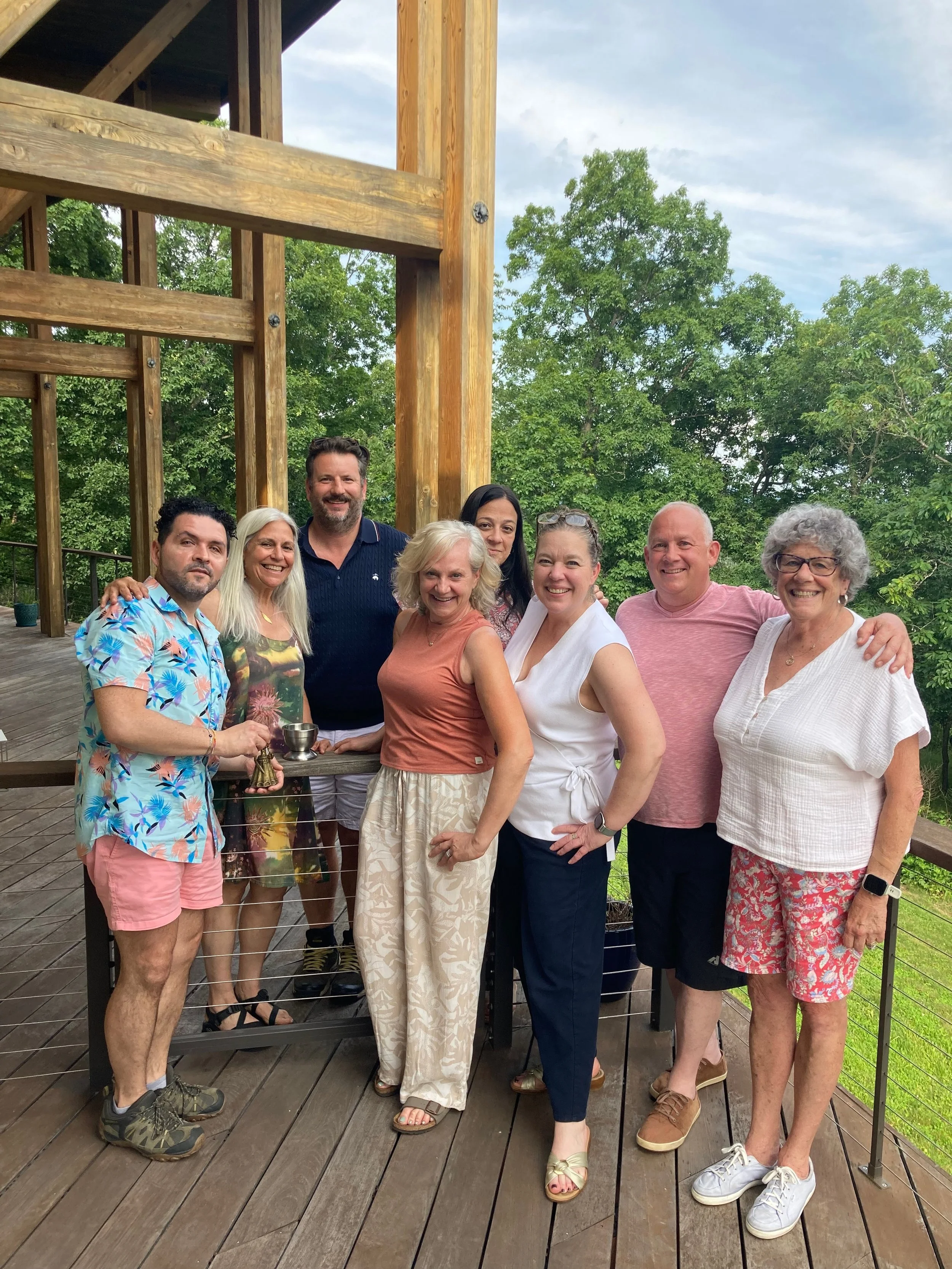 A group of nine people smiling and posing together outdoors on a wooden deck with green trees in the background.