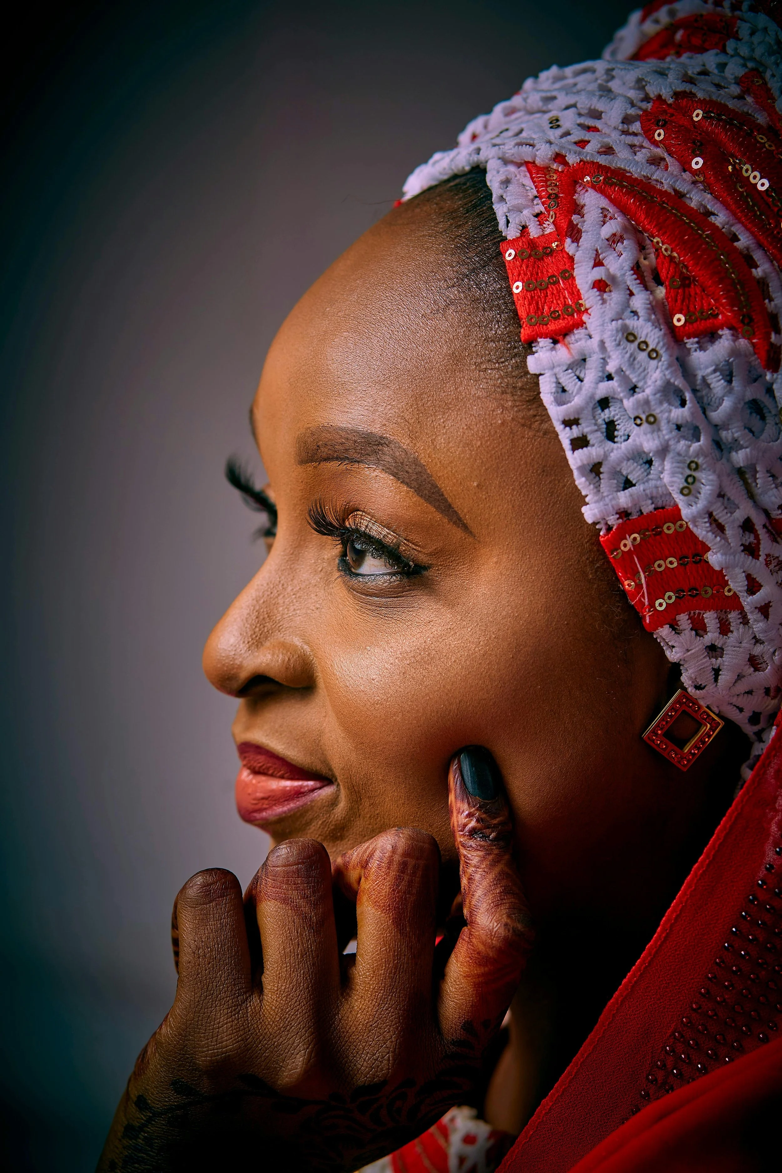 Close-up of a woman with traditional African attire, including a lace and beaded headwrap, with makeup and henna on her hand, looking thoughtfully to the side.