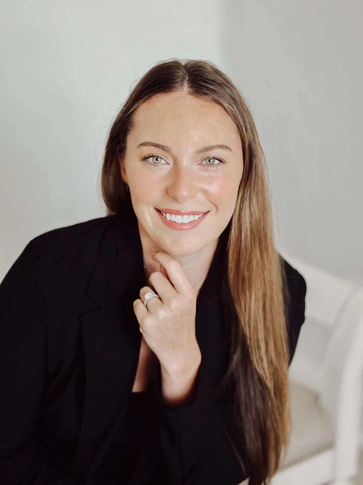 A woman with long wavy brown hair and a blue shirt sitting on a tan sofa with white and striped pillows in a living room, smiling at the camera.