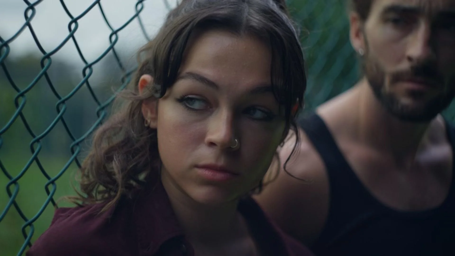 A young woman with dark hair and a nose ring, sitting behind a chain-link fence with a man partially visible beside her, outdoors during daytime.