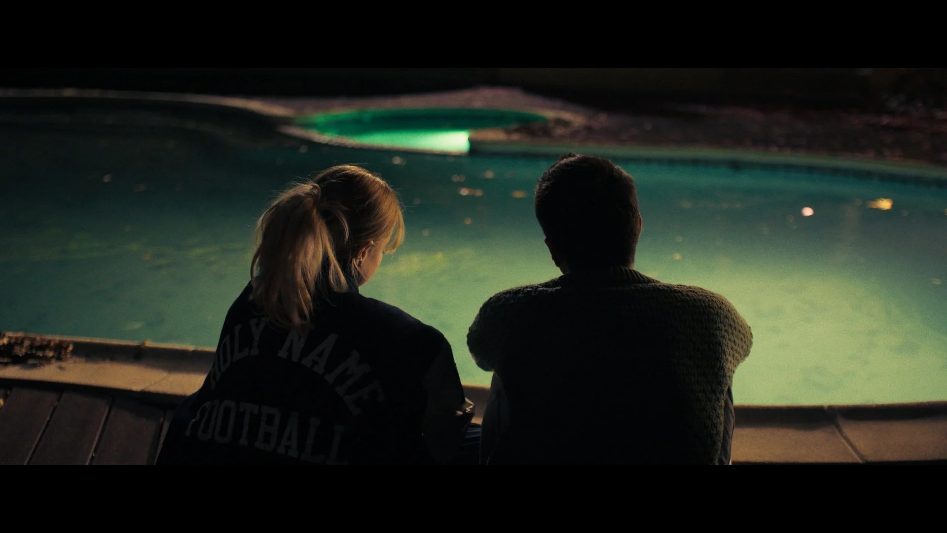 A man and woman sitting by a swimming pool at night, facing away from the camera, with their heads close together, illuminated by pool lights.