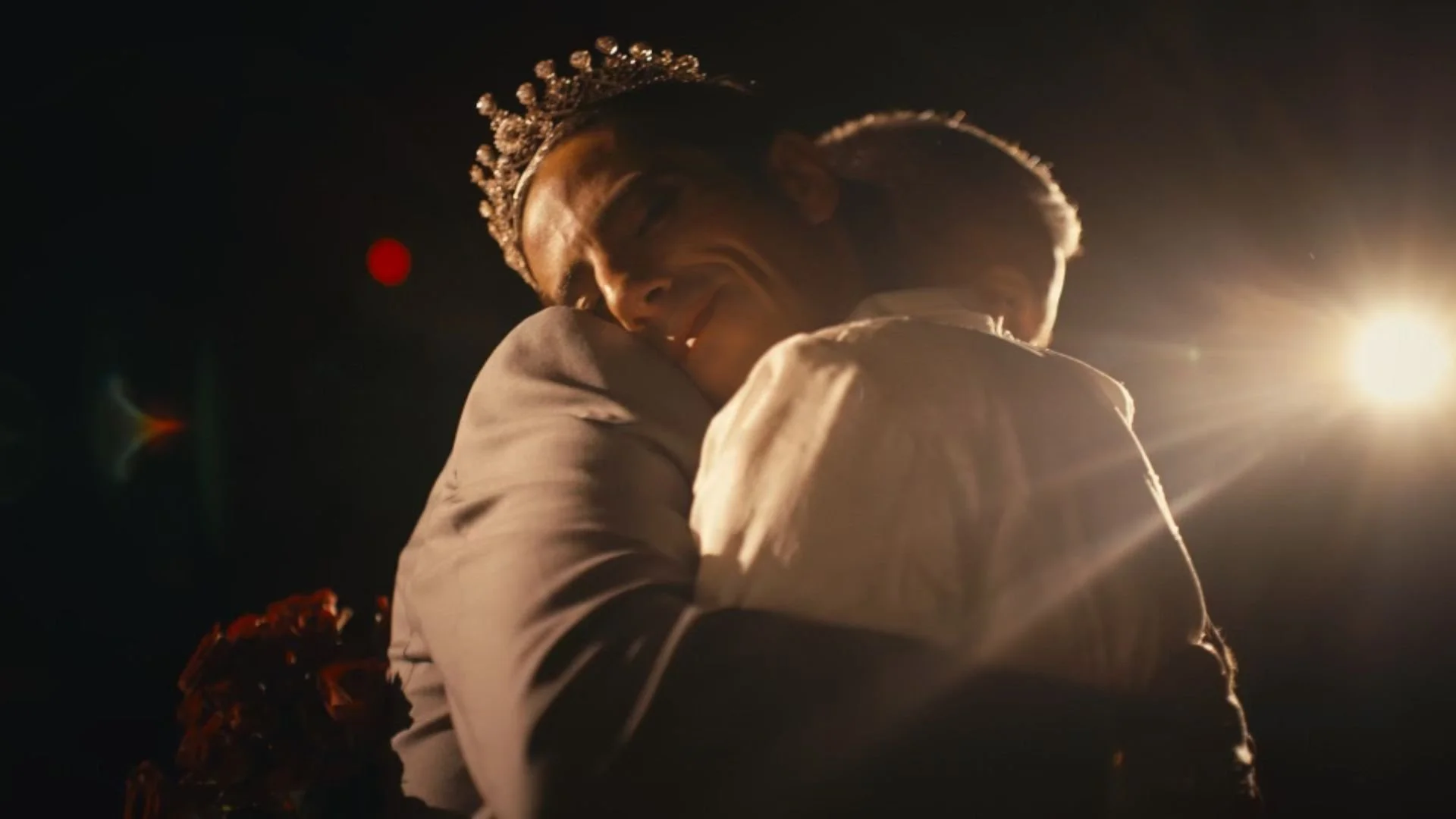 A couple hugging, the woman wearing a crown, in a dark setting with bright light shining behind them.