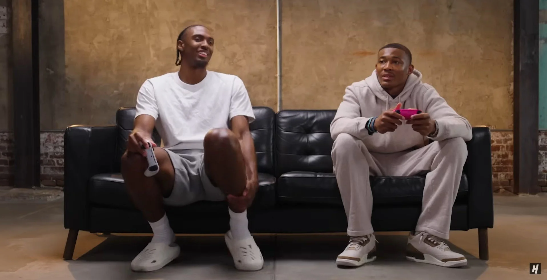 Two young men sitting on a black leather couch, playing video games in a room with brown walls and a brick section, one wearing a white t-shirt and shorts, holding a game controller, the other in a hoodie and sweatpants, also holding a game controlle