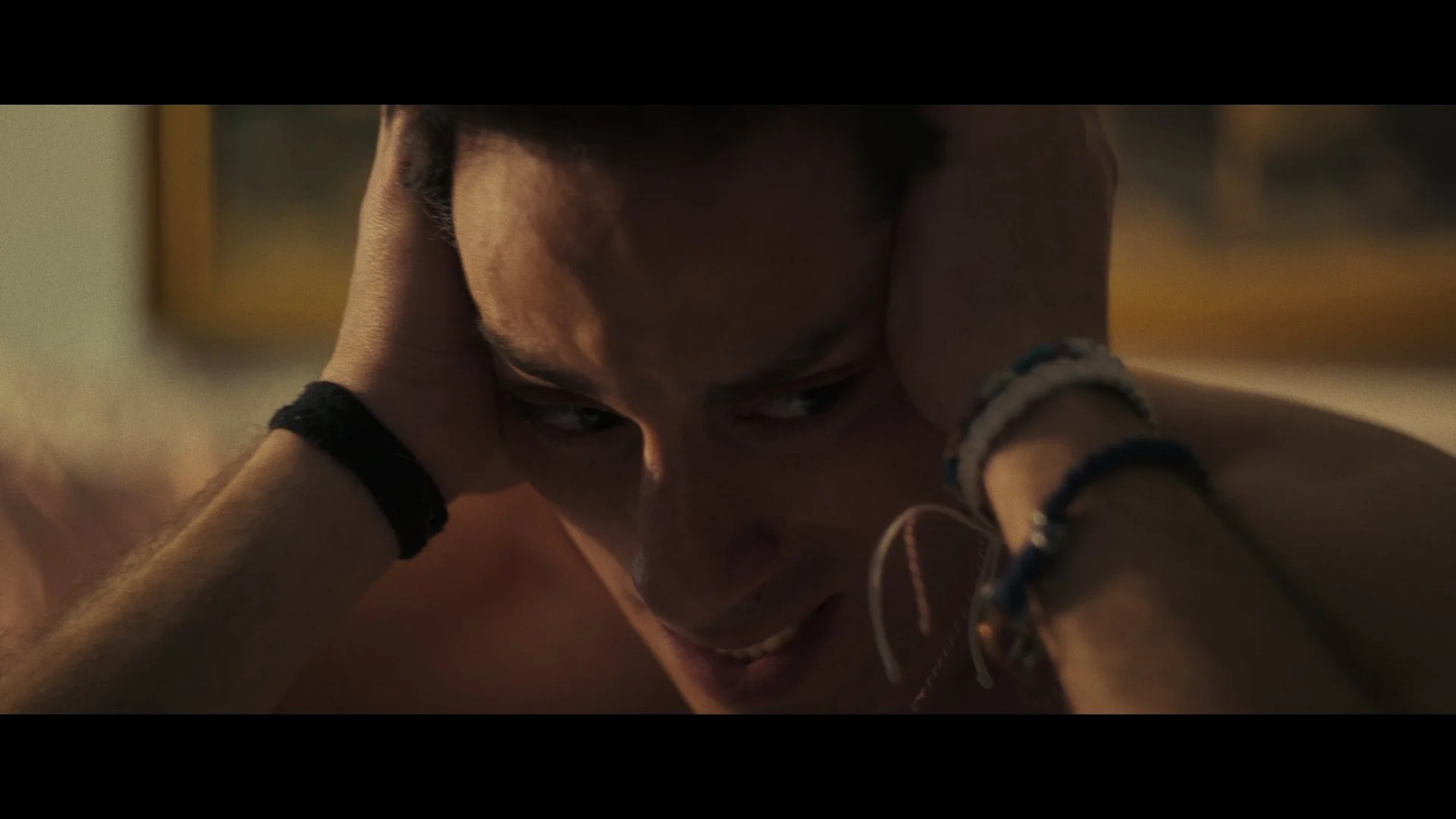 A shirtless young man with dark hair and multiple bracelets on his wrists, holding his head with an expression of distress or frustration.