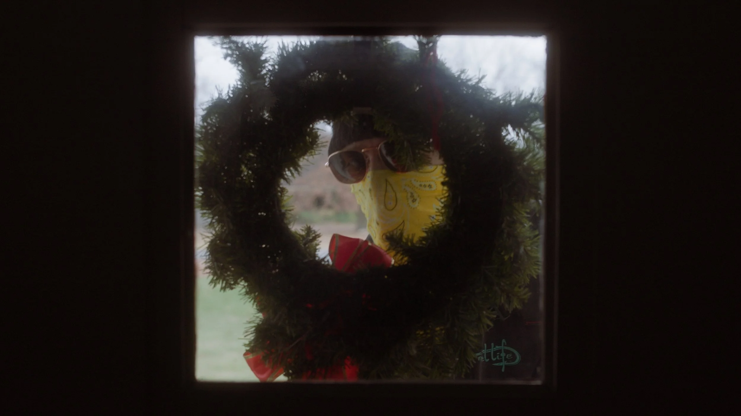 Person wearing sunglasses, a yellow bandana, and a red shirt, looking through a Christmas wreath on a door window.