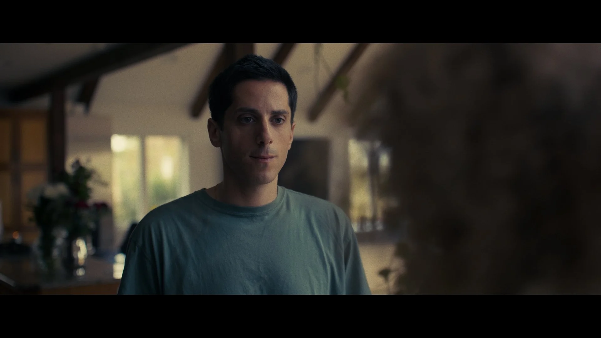 A man with short dark hair looking at a large, blurred object in a warmly lit room with large windows and wooden ceiling beams.