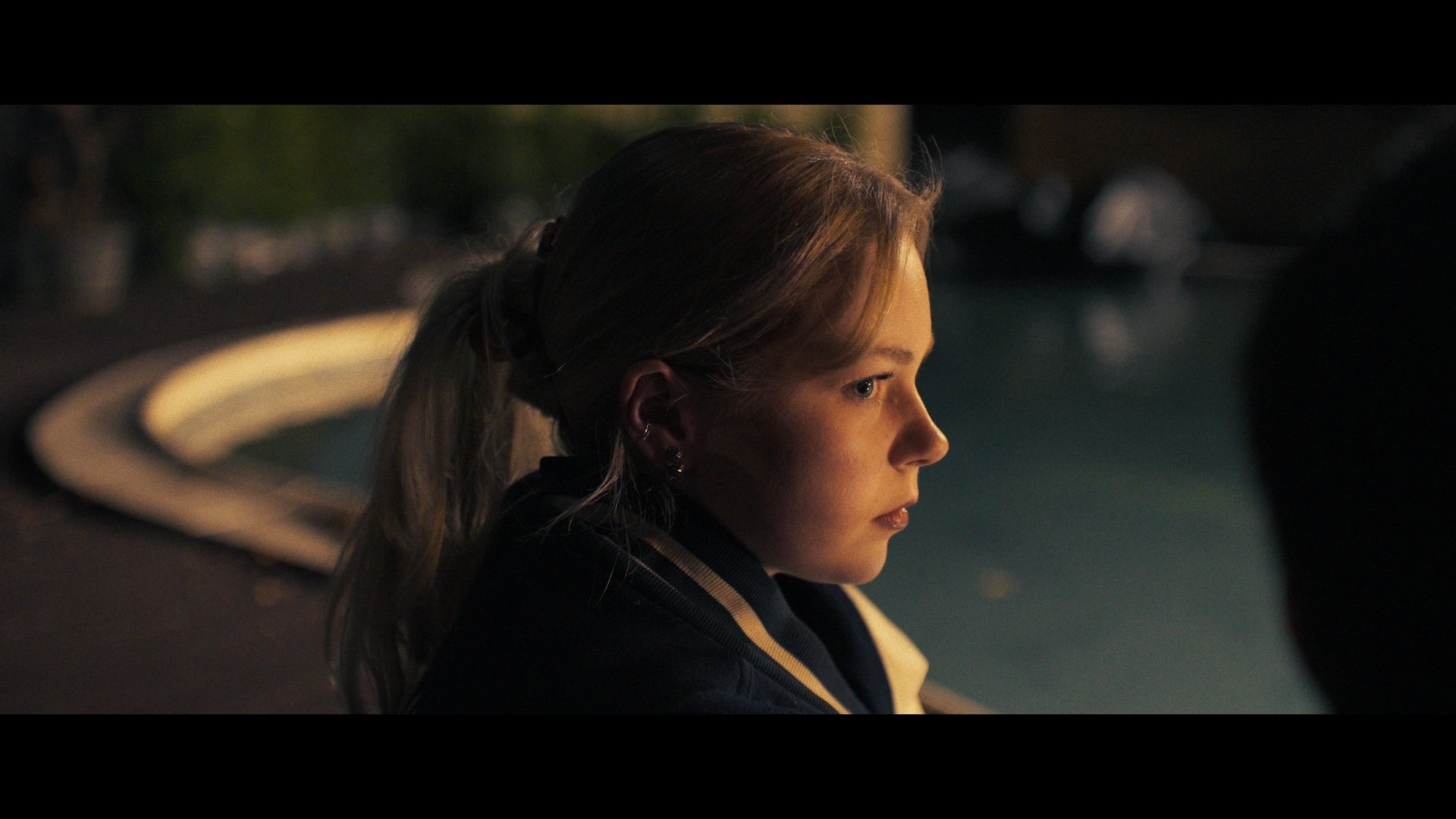 Side profile of a young woman with blonde hair, styled in a ponytail, looking thoughtfully to the right at nighttime, in front of a fountain with blurred lights in the background.