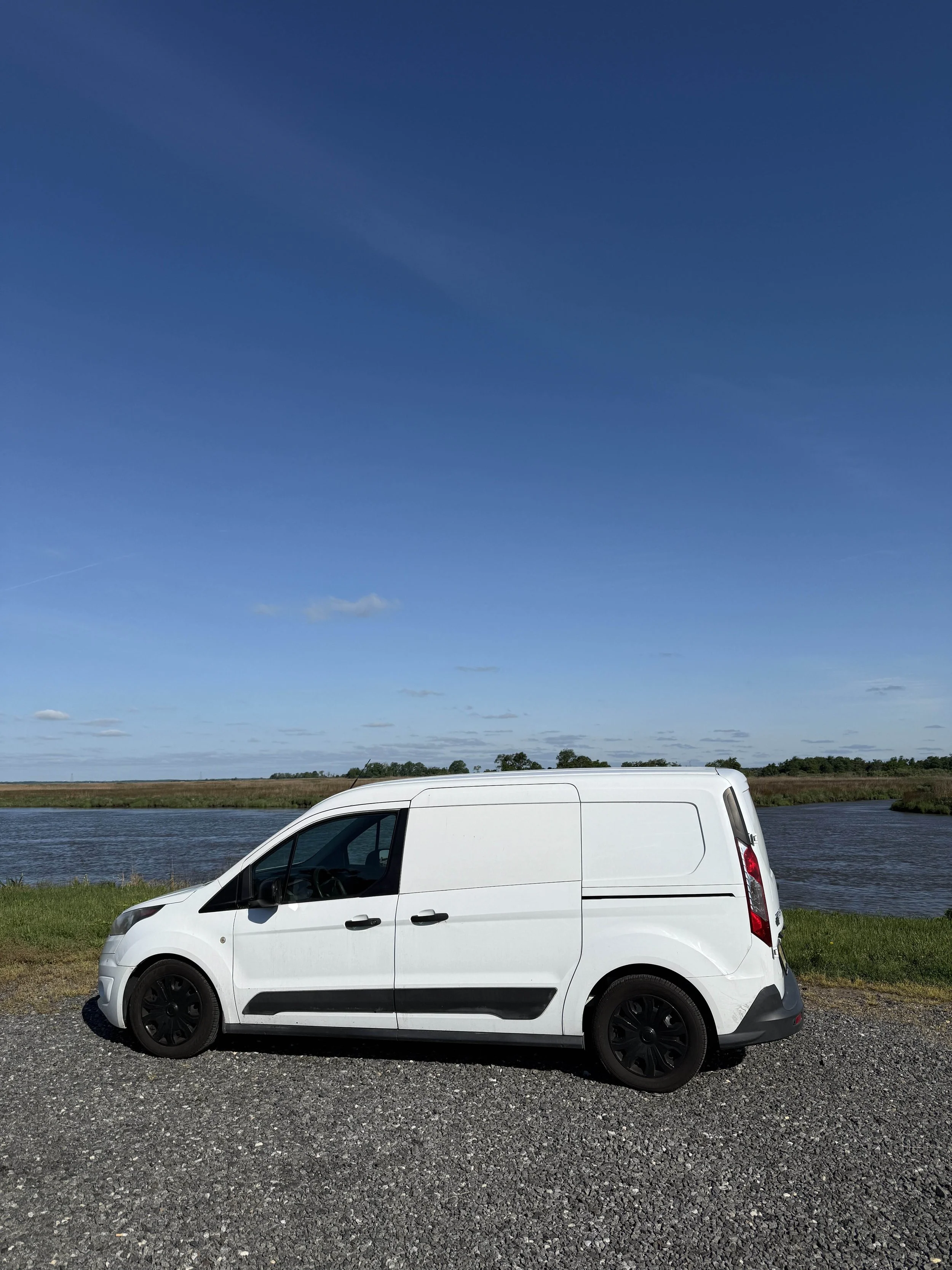White van parked on gravel at a lakeside with grass and water, under a clear blue sky.
