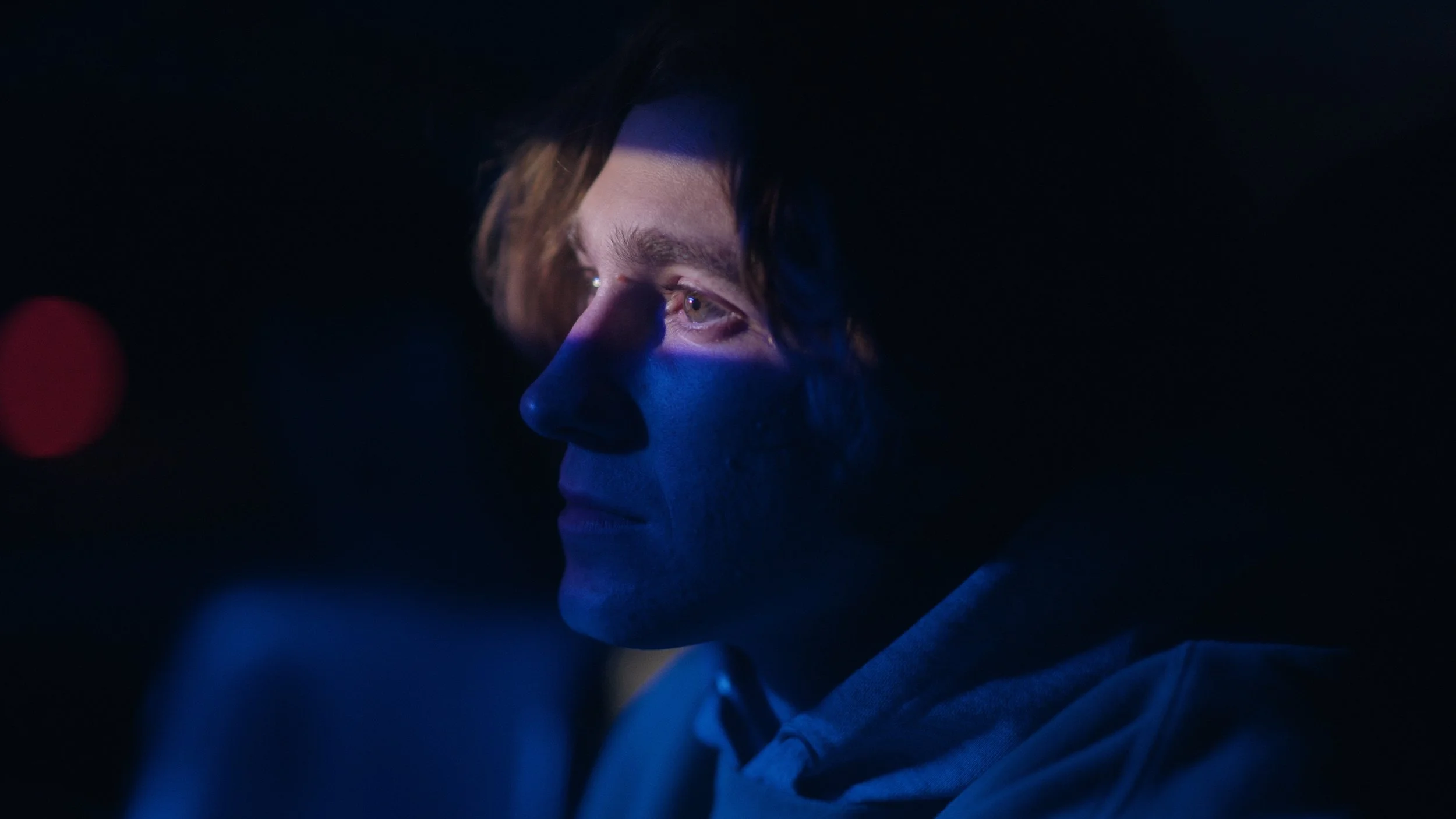 A young man with light brown hair looking into the distance at night, illuminated by a blue light.