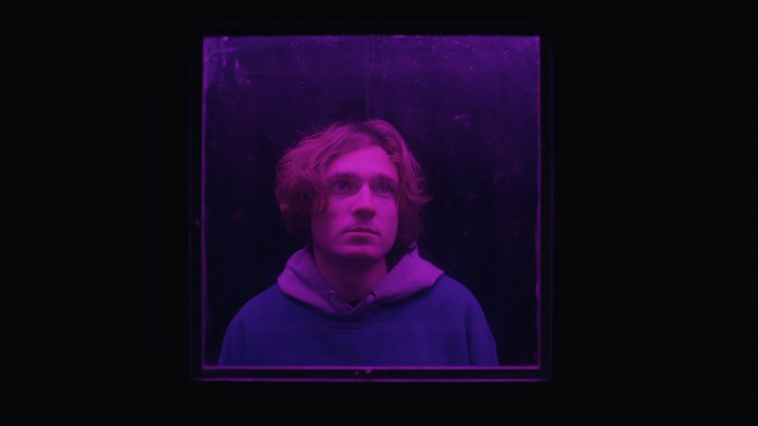 Young man with wavy hair wearing a hoodie, looking upwards, illuminated by purple light inside a glass box.