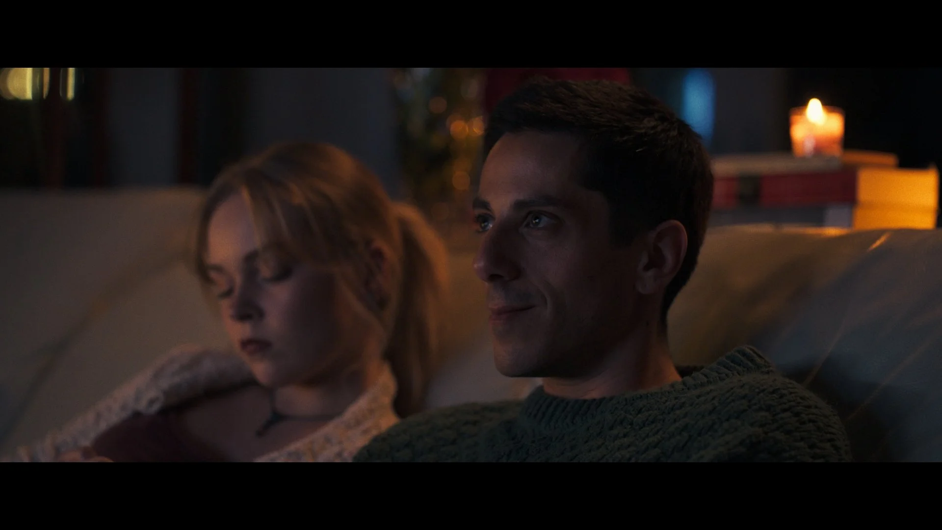A man and woman sitting on a couch in a dimly lit room with a lit candle and books in the background. The man is smiling while the woman has her eyes closed.