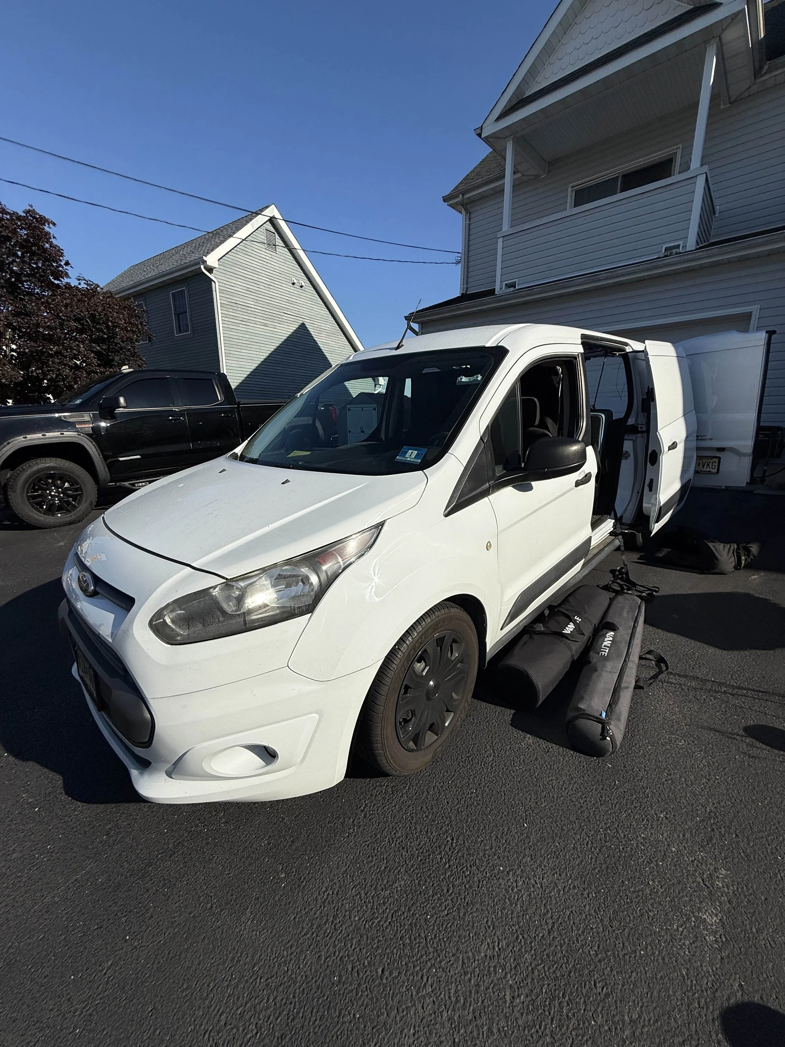 White cargo van parked on driveway with open side door, black side mirror, and black vehicle parked next to it, in front of residential houses on a clear, sunny day.