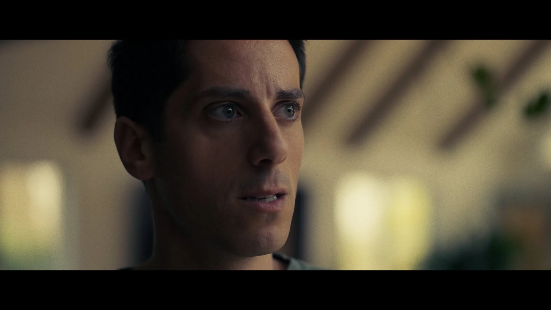 Close-up of a young man with blue eyes and short dark hair, looking serious, indoor setting with blurred background.