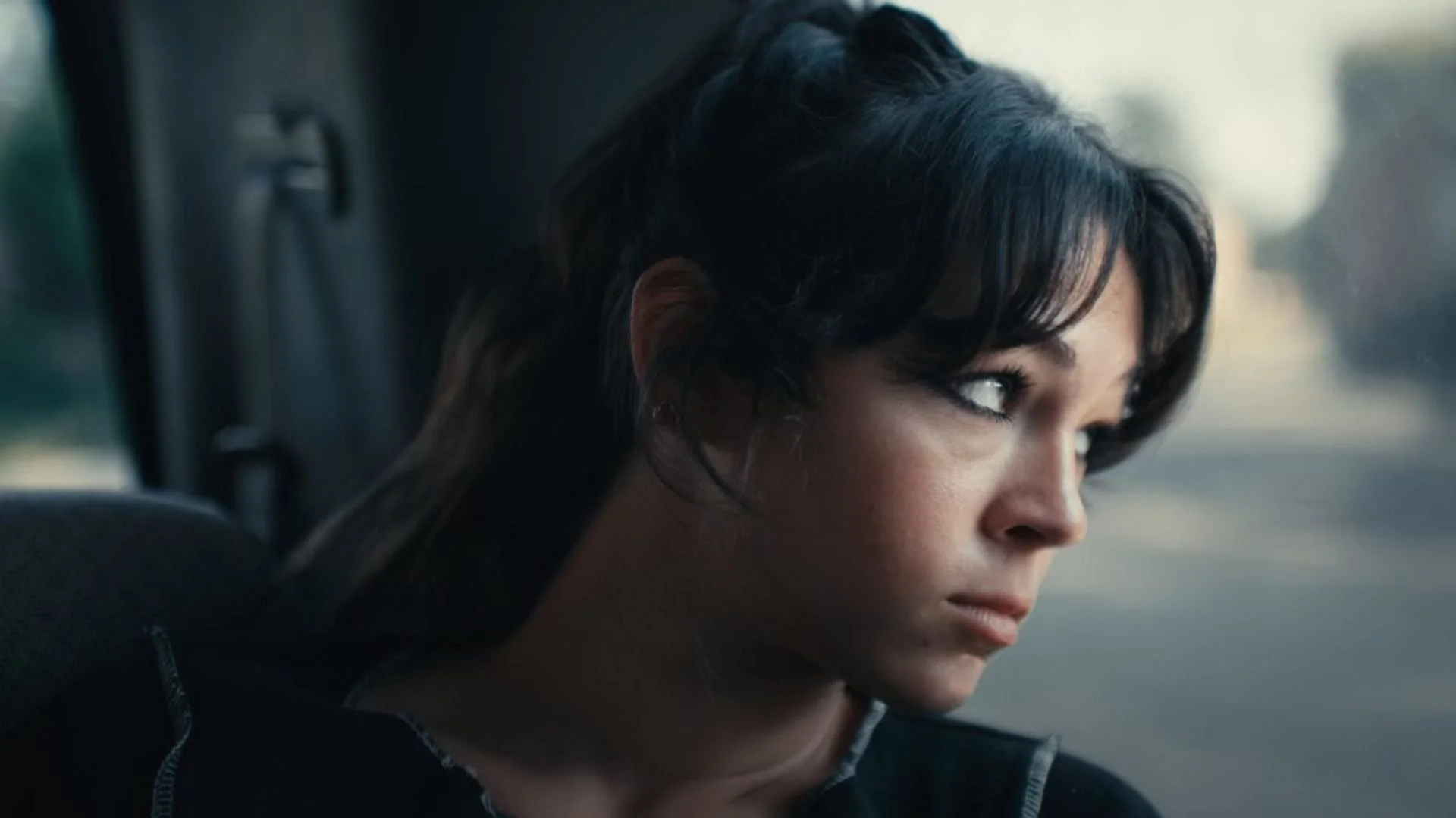A woman with short dark hair looking out of a window with a serious expression.
