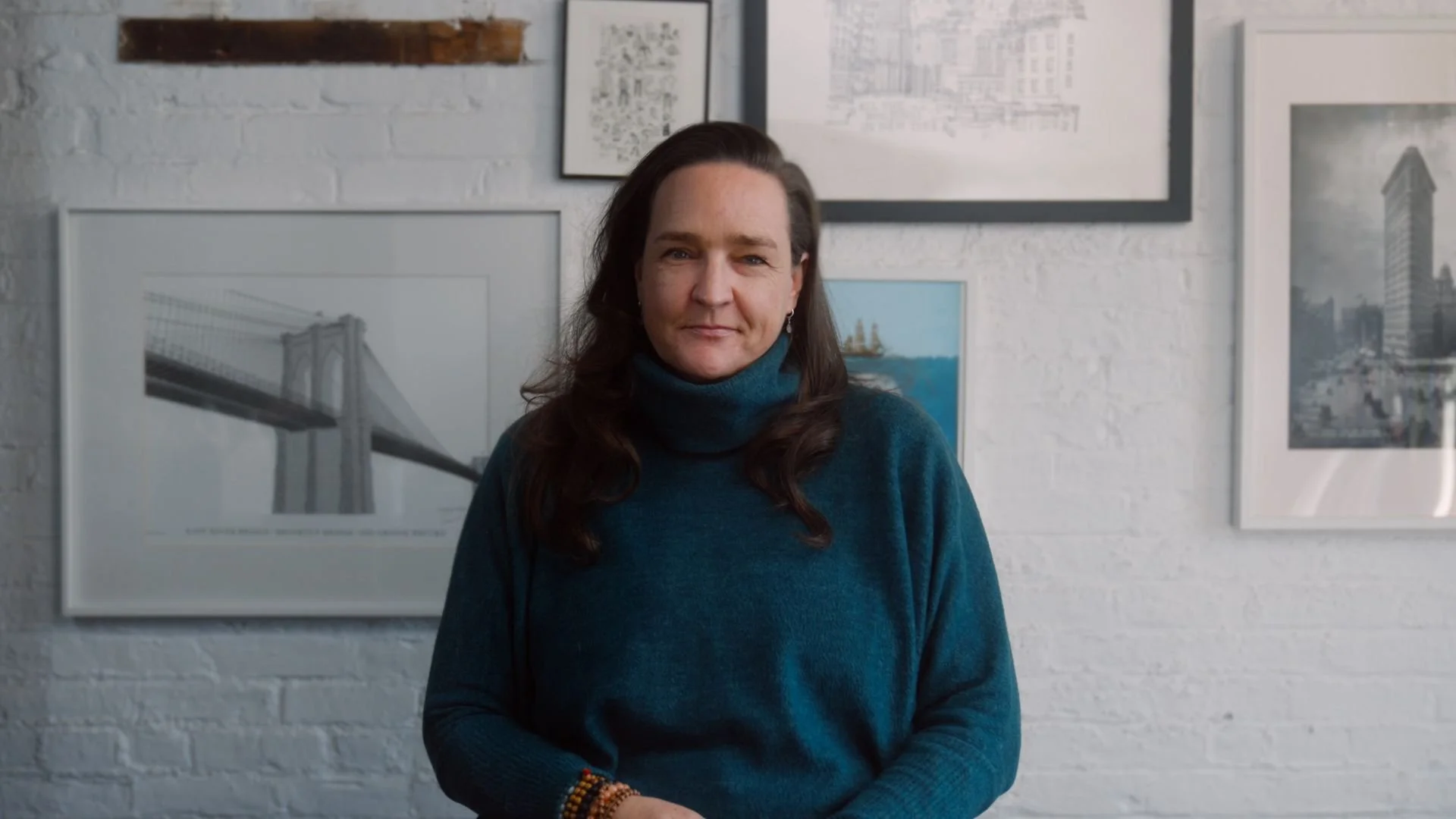 A woman with long dark hair wearing a blue turtleneck sweater standing in front of a white brick wall with framed artwork, including a bridge illustration and architectural photos.