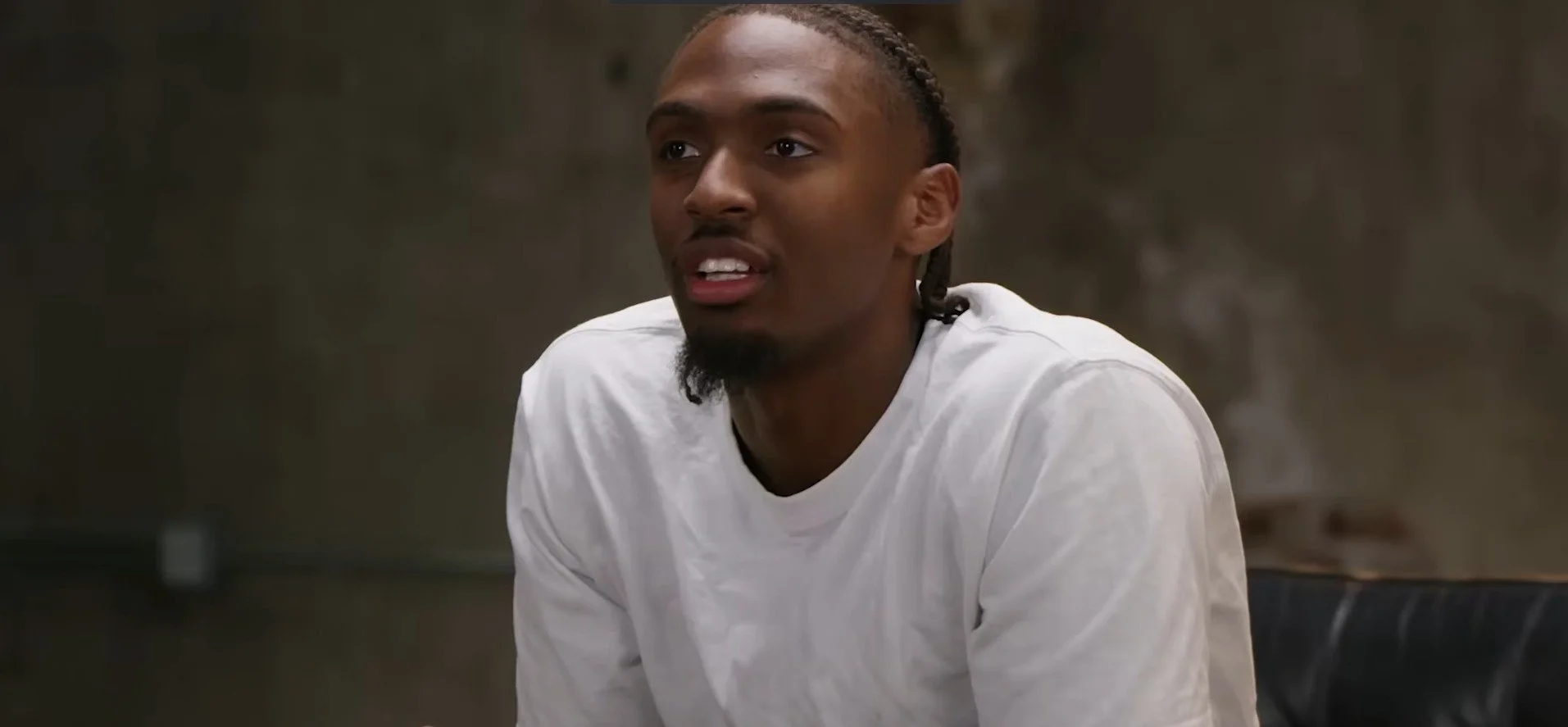 A young man with braided hair and a goatee, wearing a white shirt, sitting and looking slightly to his left in an indoor setting.