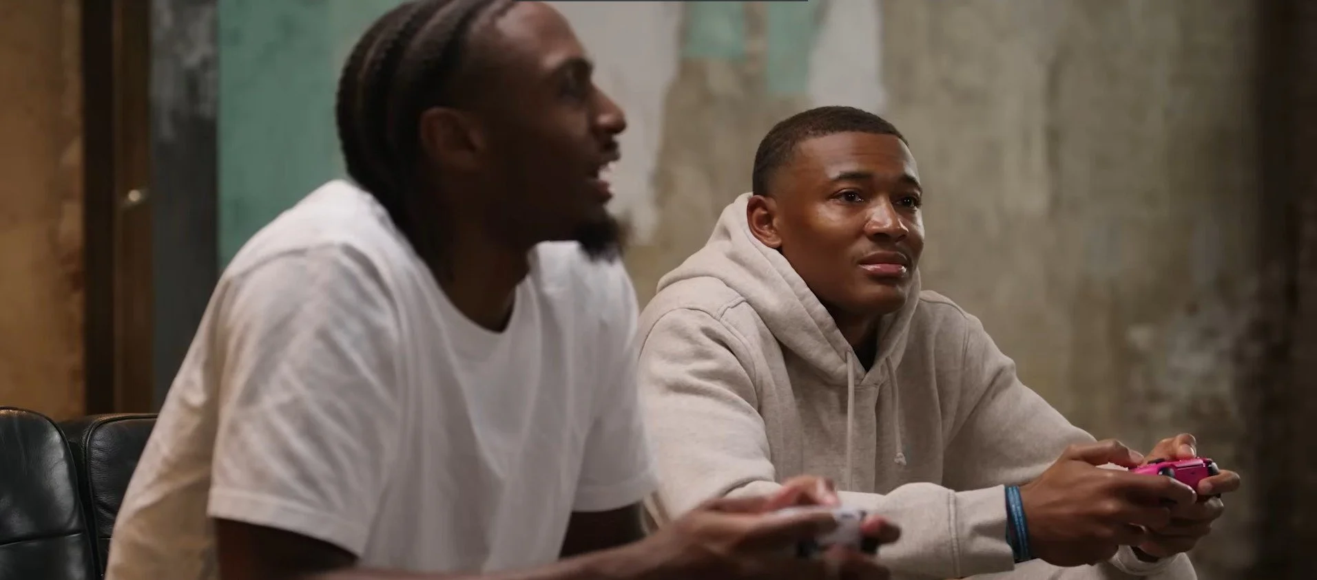 Two young men sit side by side playing video games with controllers, one wearing a white t-shirt and the other in a beige hoodie, in a room with rustic walls.