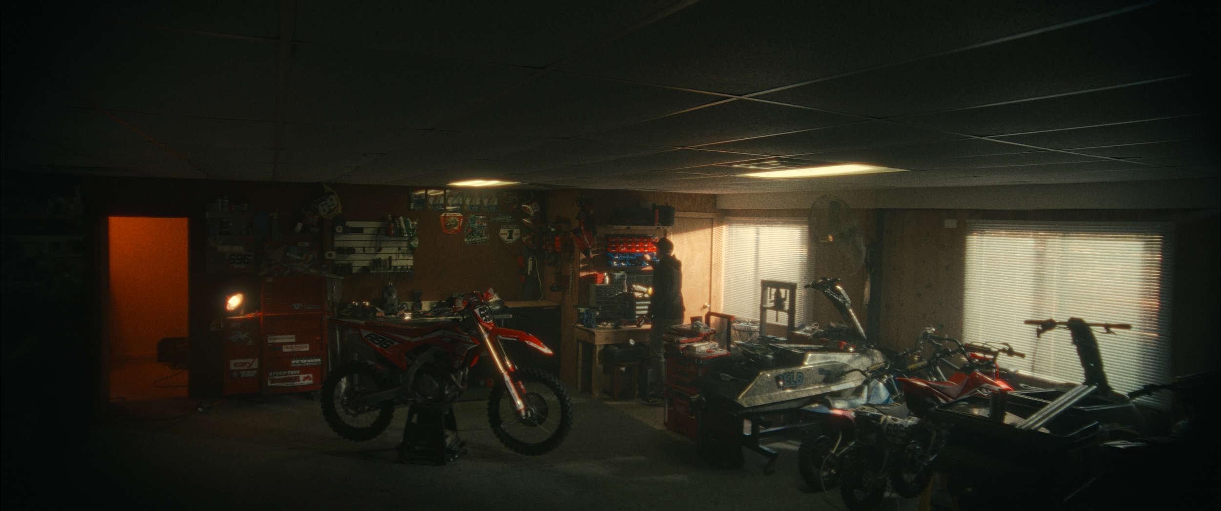 A dimly lit garage with a person working at a workbench surrounded by motorcycles, tools, and equipment, with windows letting in natural light.