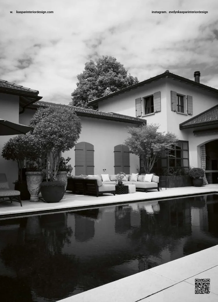 A backyard with a swimming pool, outdoor seating area with cushions, potted plants, and trees in front of a house with shuttered windows and a tiled roof, under a cloudy sky.